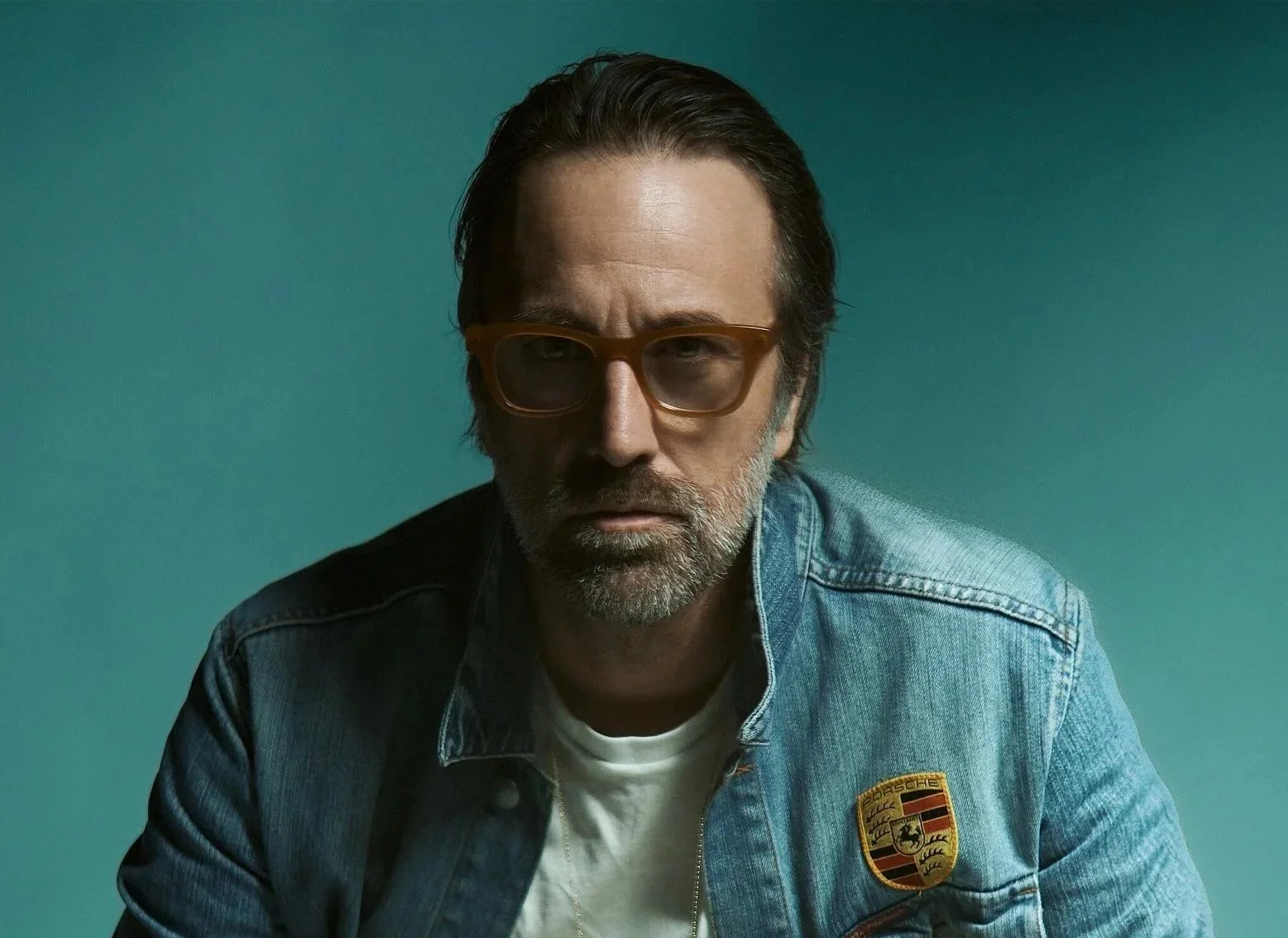 Man with dark hair, beard, and glasses wearing a denim jacket with Porsche patch, sitting against teal background.