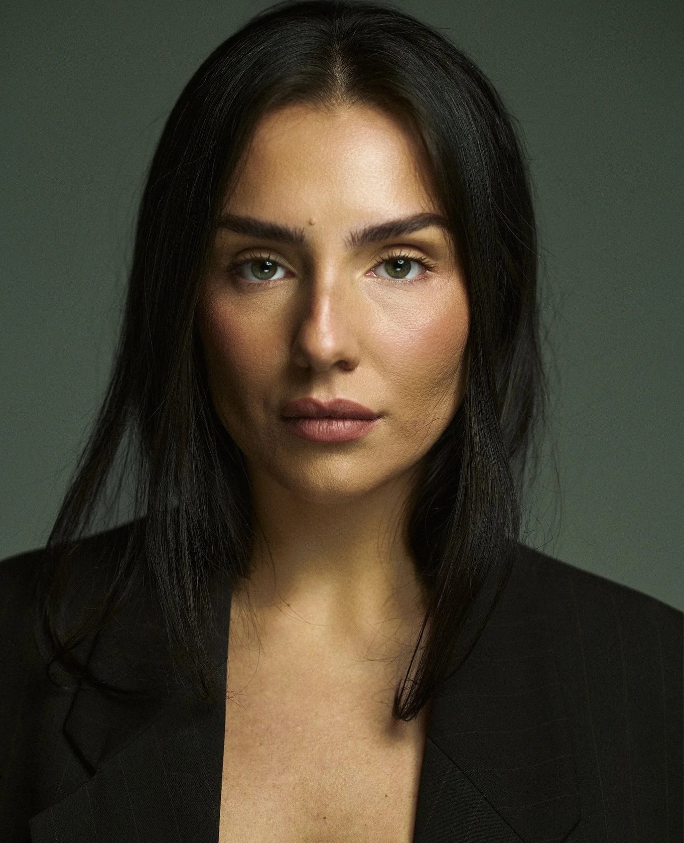 A woman with long, dark hair and blue eyes wearing a black blazer against a plain background.