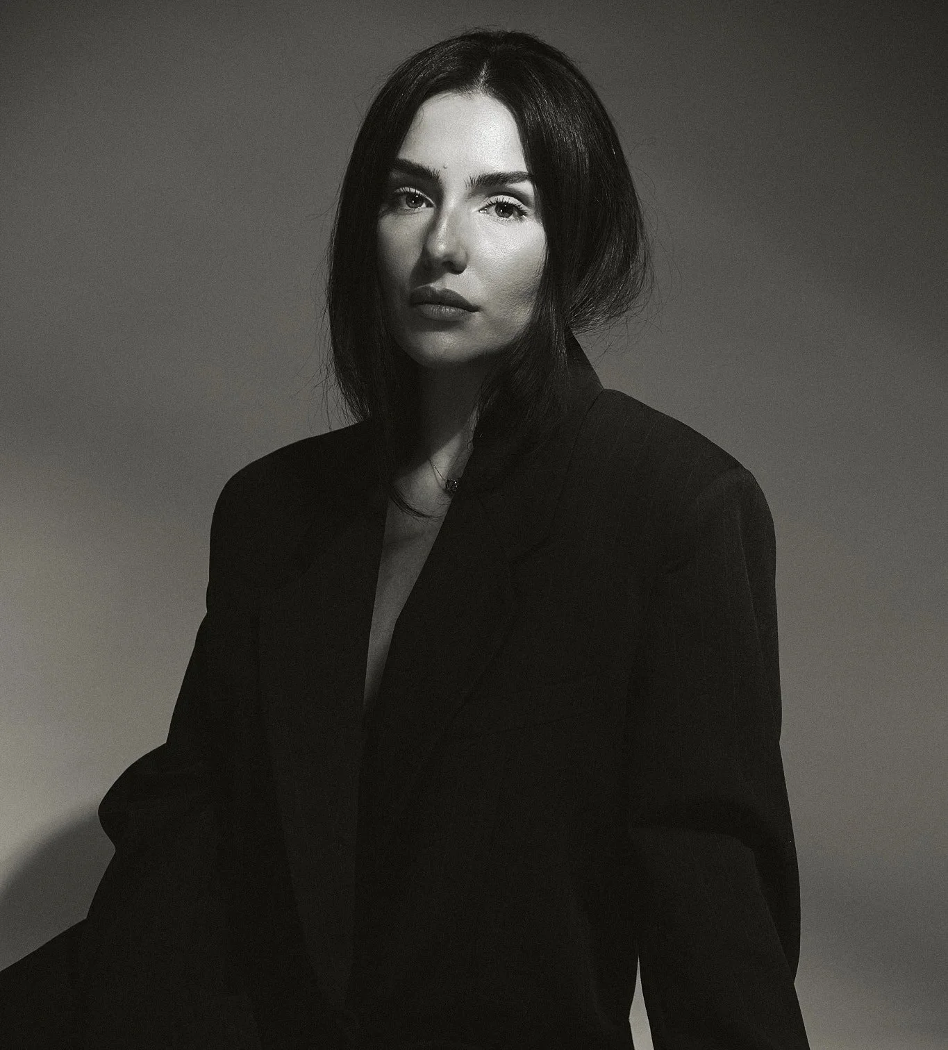 A black-and-white portrait of a woman with dark hair, wearing a dark blazer, looking directly at the camera with a serious expression.