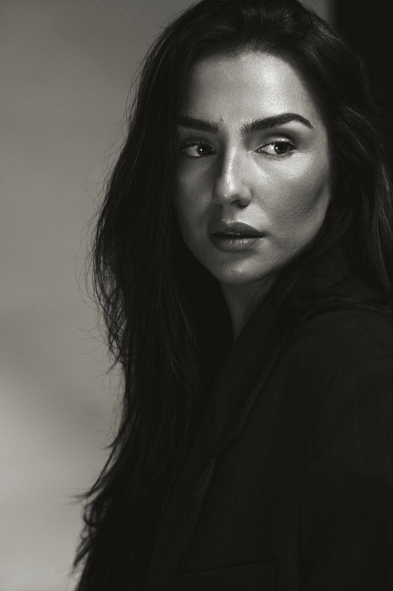 A black and white close-up portrait of a woman with long, dark hair and makeup, looking slightly to the side.