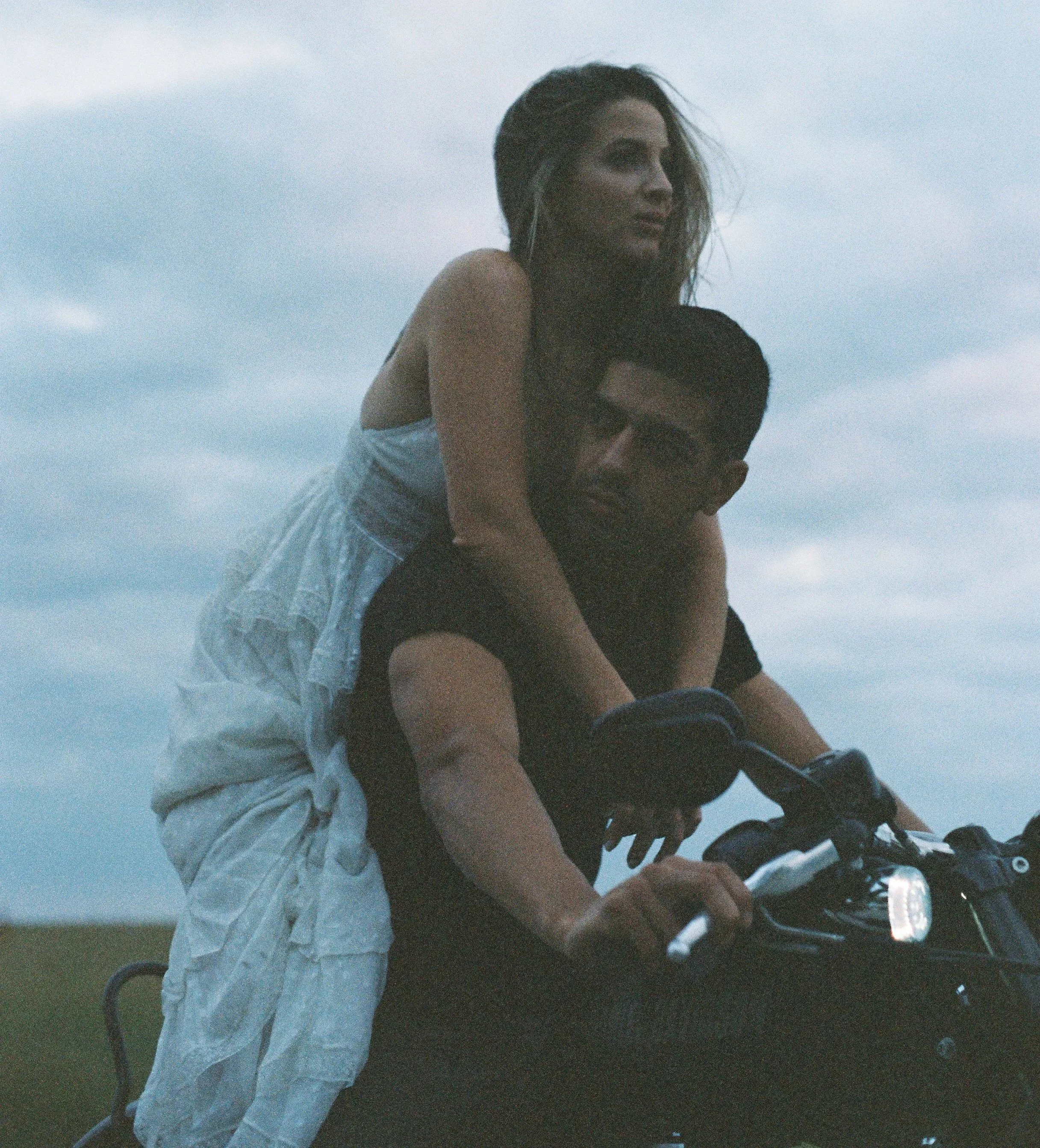 A woman with long brown hair, wearing a black top and light-colored pants, riding a motorcycle with an open sky background.
