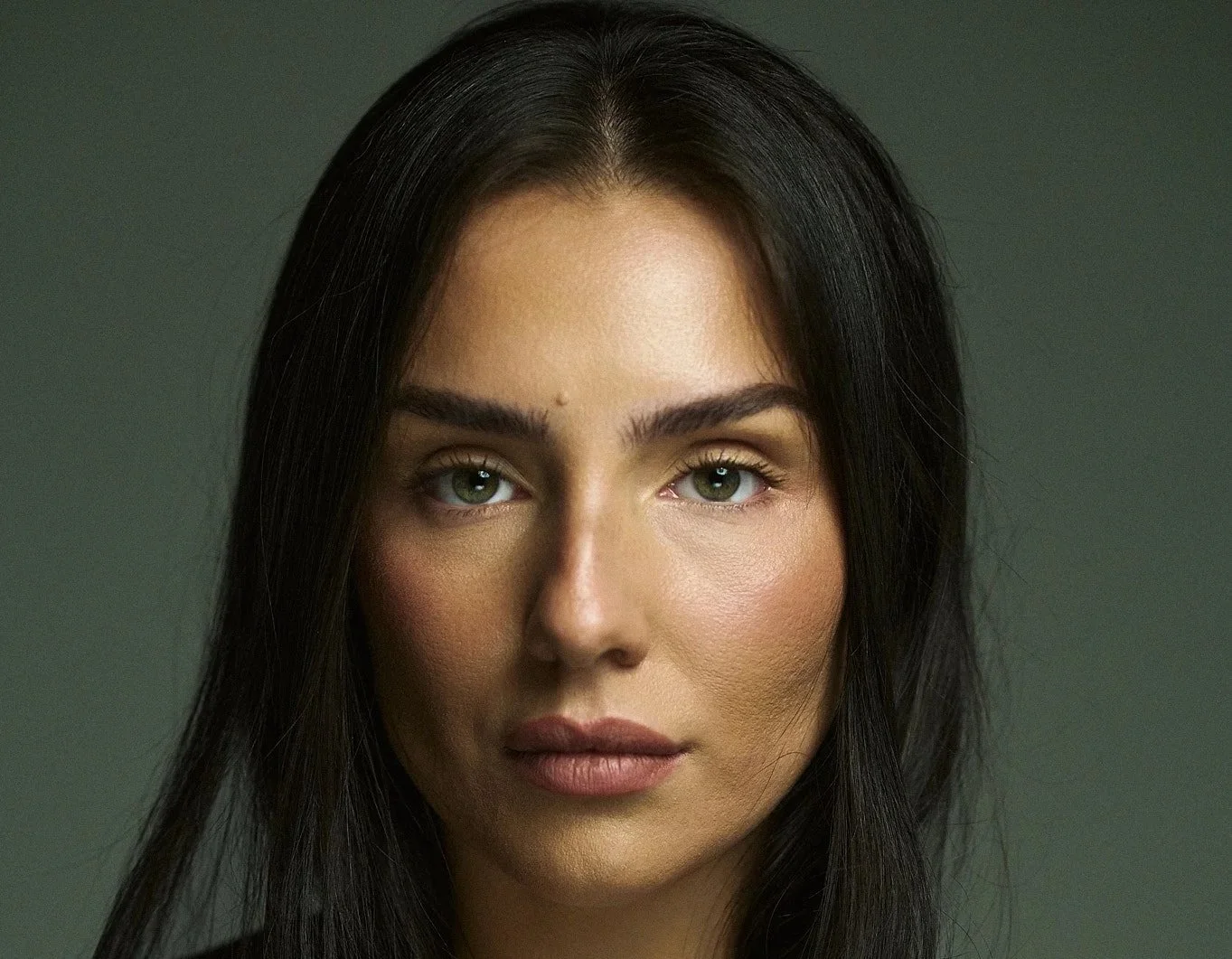 Close-up portrait of a woman with long dark hair and green eyes, looking directly at the camera against a plain dark background.