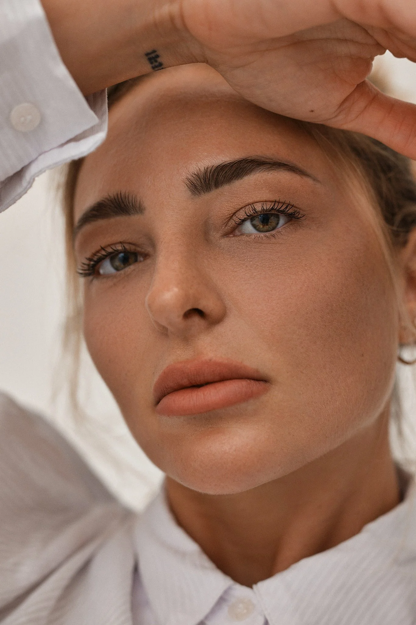 Close-up of a woman lying down, with her hand resting on her forehead. She has well-defined eyebrows, long eyelashes, and smooth skin, wearing a white shirt with a collar.