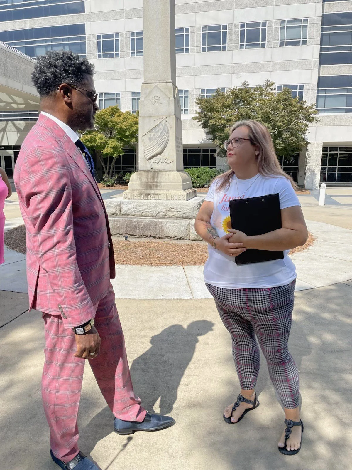 A man in a pink plaid suit and a woman in plaid pants are talking outside in front of a monument with a flag in front of a courthouse signaling coming together for humanity and justice.