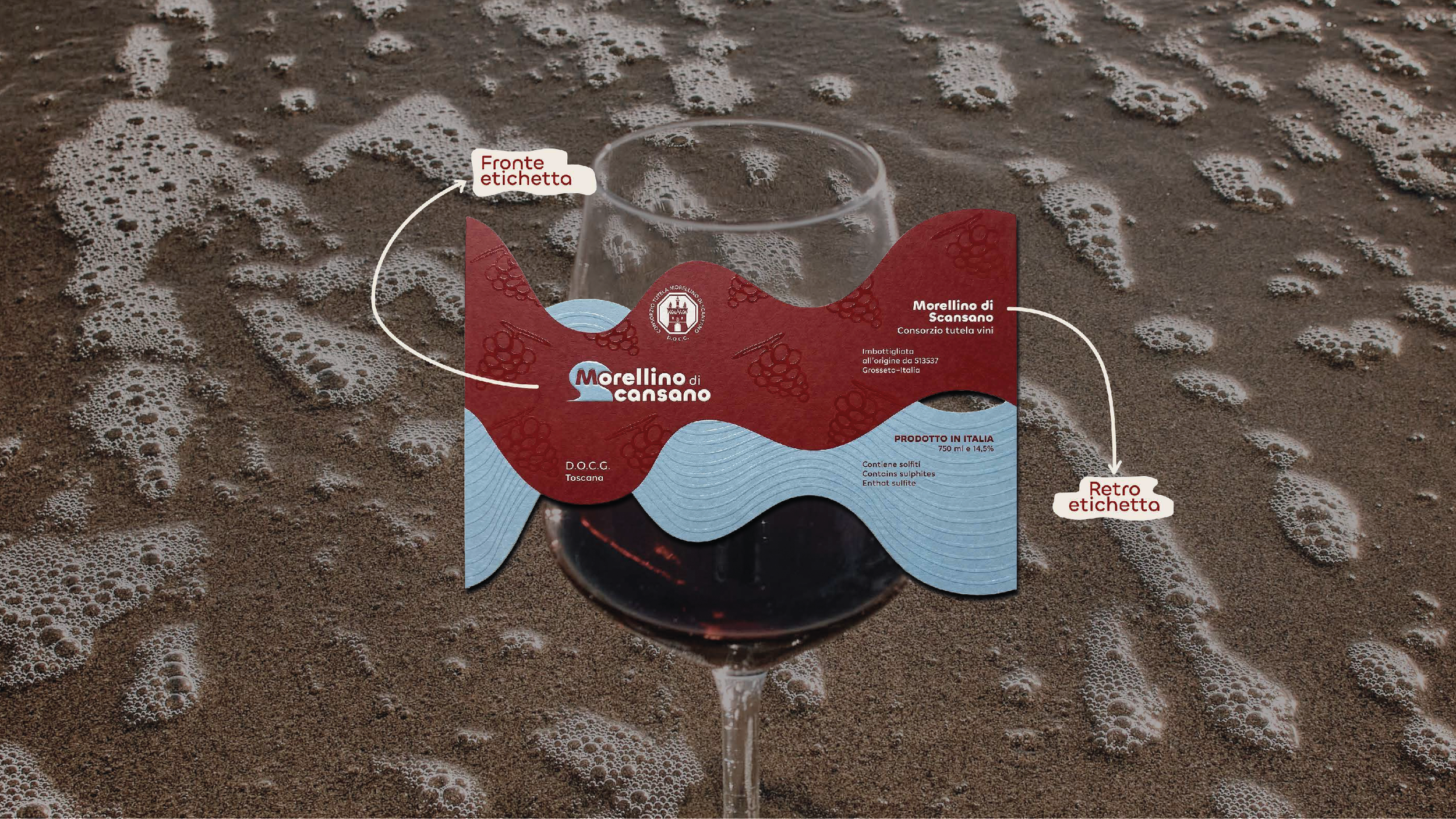 Etichetta di vino Morellino di Scansano posizionata su un bicchiere di vino rosso, circondato da schiuma e acqua sulla spiaggia