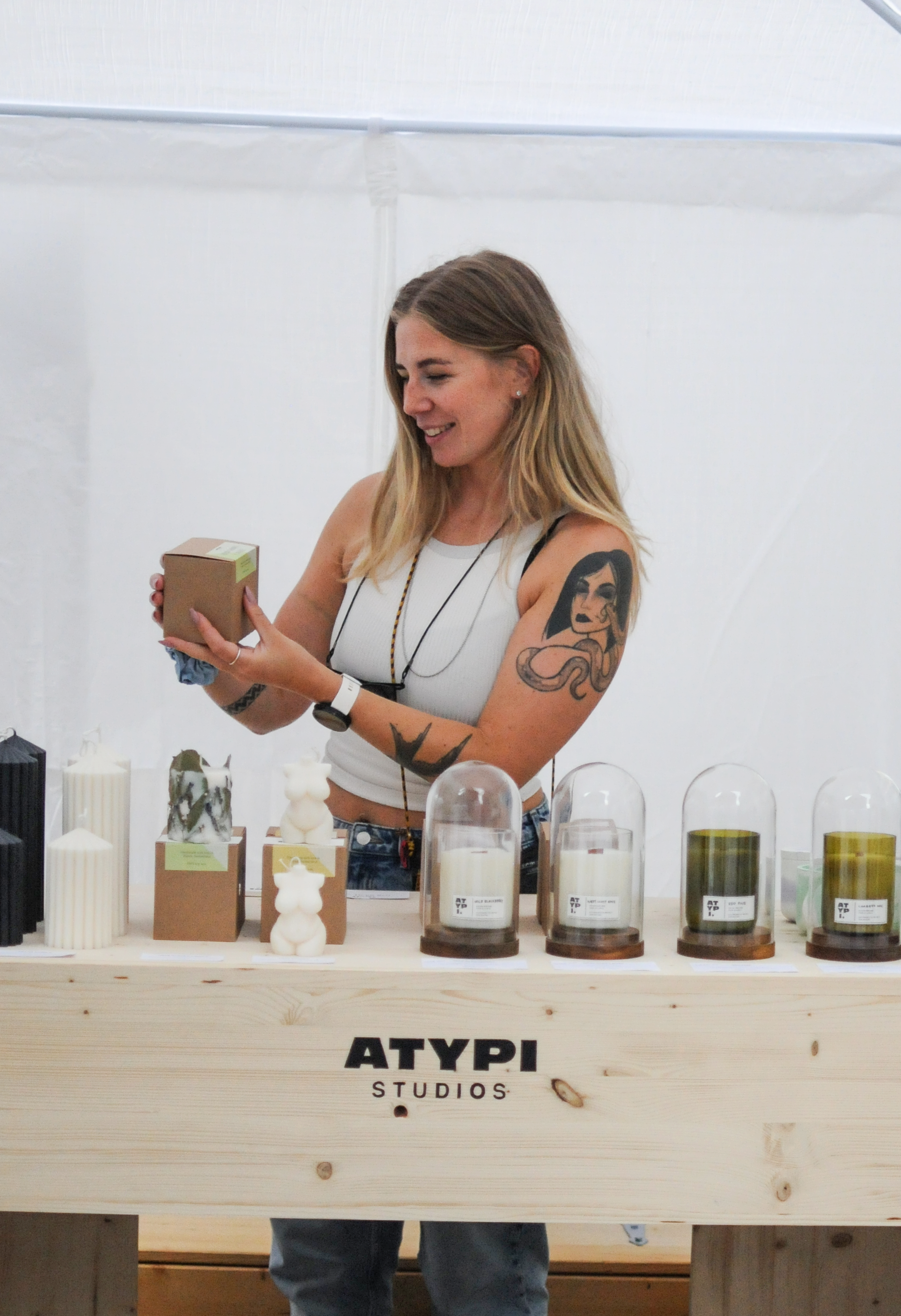 A woman with shoulder-length hair, tattoos on her arms, and wearing a white tank top is smiling and looking down while holding a small cardboard box. She stands behind a wooden table displaying various candles and decorative items. The table has the words 'ATYPI STUDIOS' printed on its front.