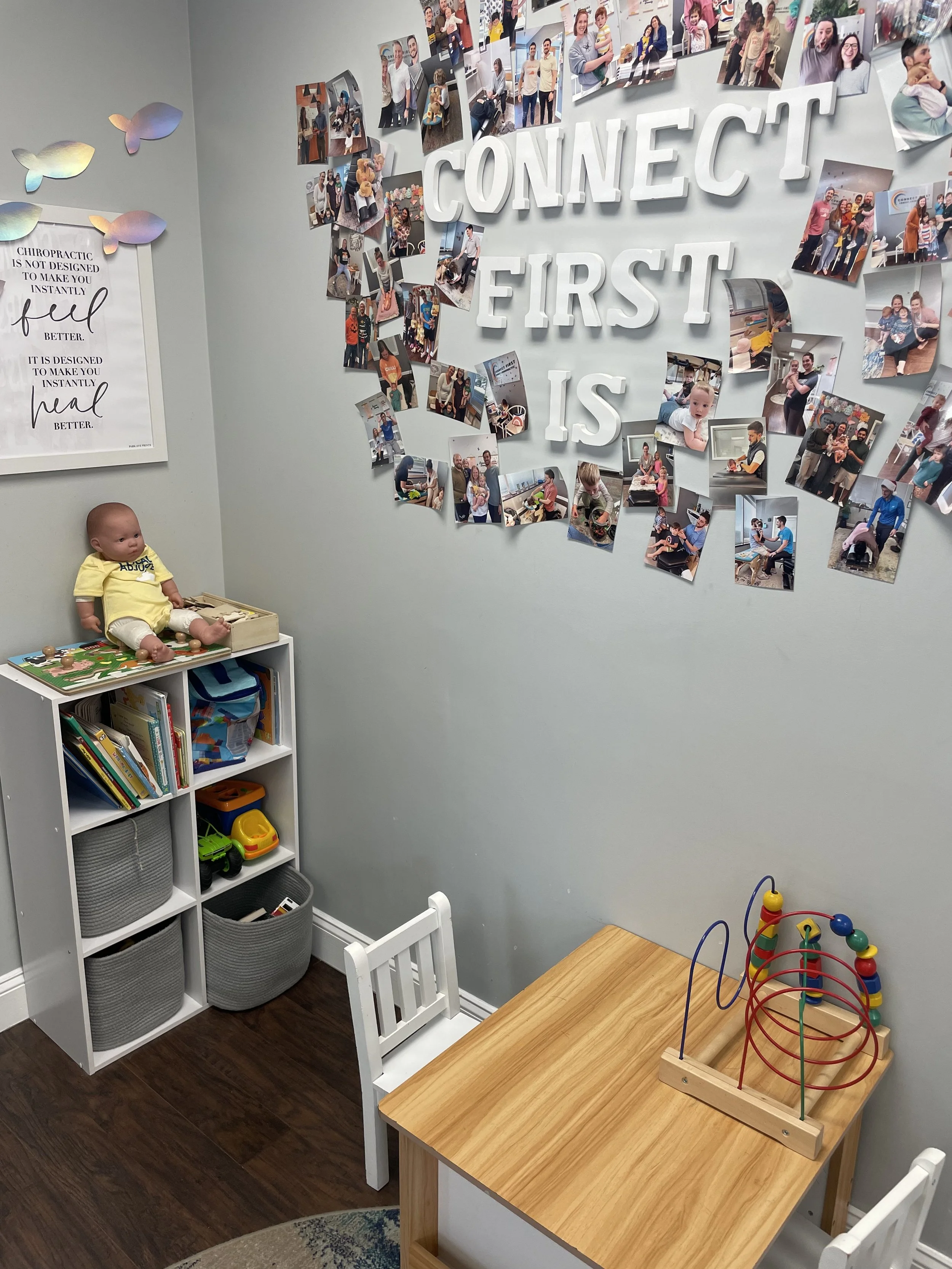 A children's playroom with a wall decorated with a collage of photos and large white letters spelling 'Connect First.' A doll is seated on a small white bookshelf filled with books and bins, and there is a wooden table with a bead maze toy on it. Nearby, colorful fish wall decor and a framed sign about chiropractic are visible.