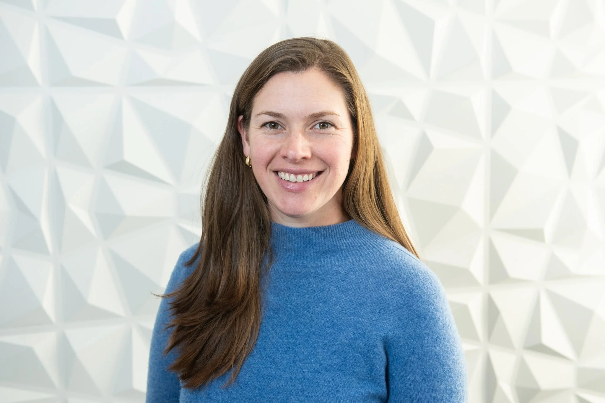 A woman with long brown hair wearing a blue sweater smiling at the camera, standing in front of a white textured wall with geometric patterns.