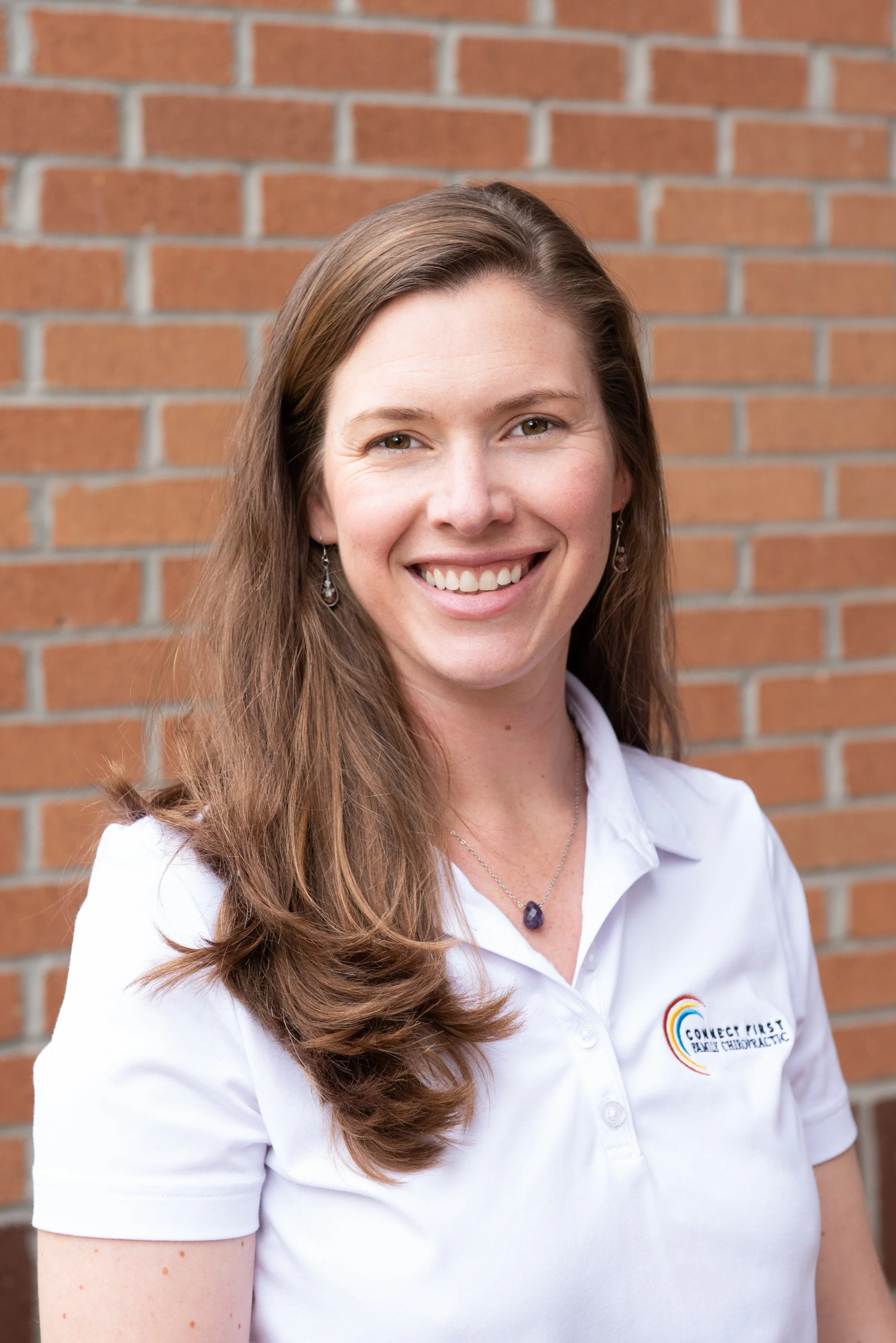 A woman with long brown hair styled in loose waves, smiling and wearing a white polo shirt with the logo "Connect First Family Chiropractic," standing in front of a brick wall, accessorized with earrings and a necklace.
