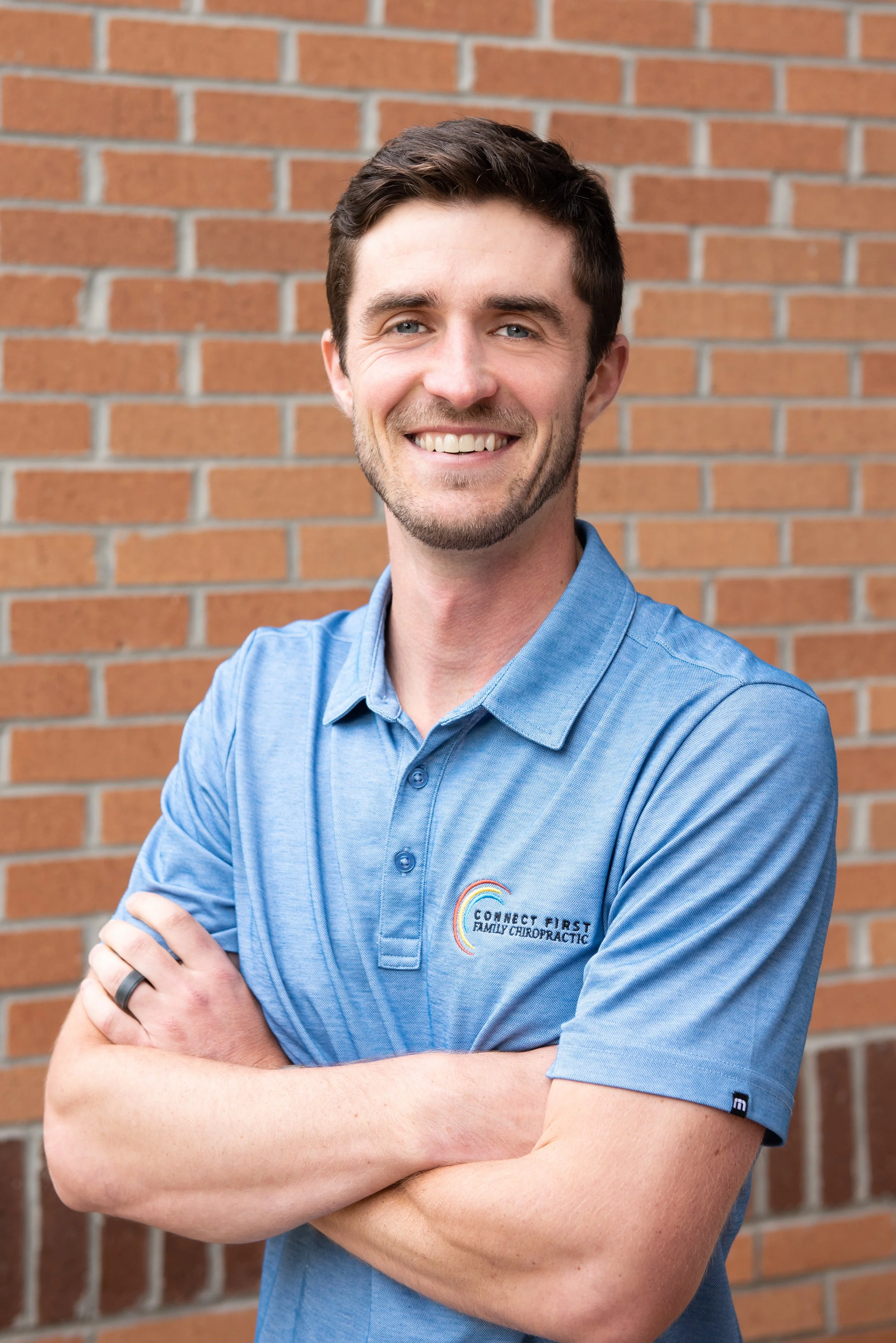 A man with short dark hair and a beard, smiling, wearing a light blue polo shirt with a logo that reads 'Connect First Family Chiropractic,' standing with arms crossed in front of a brick wall.