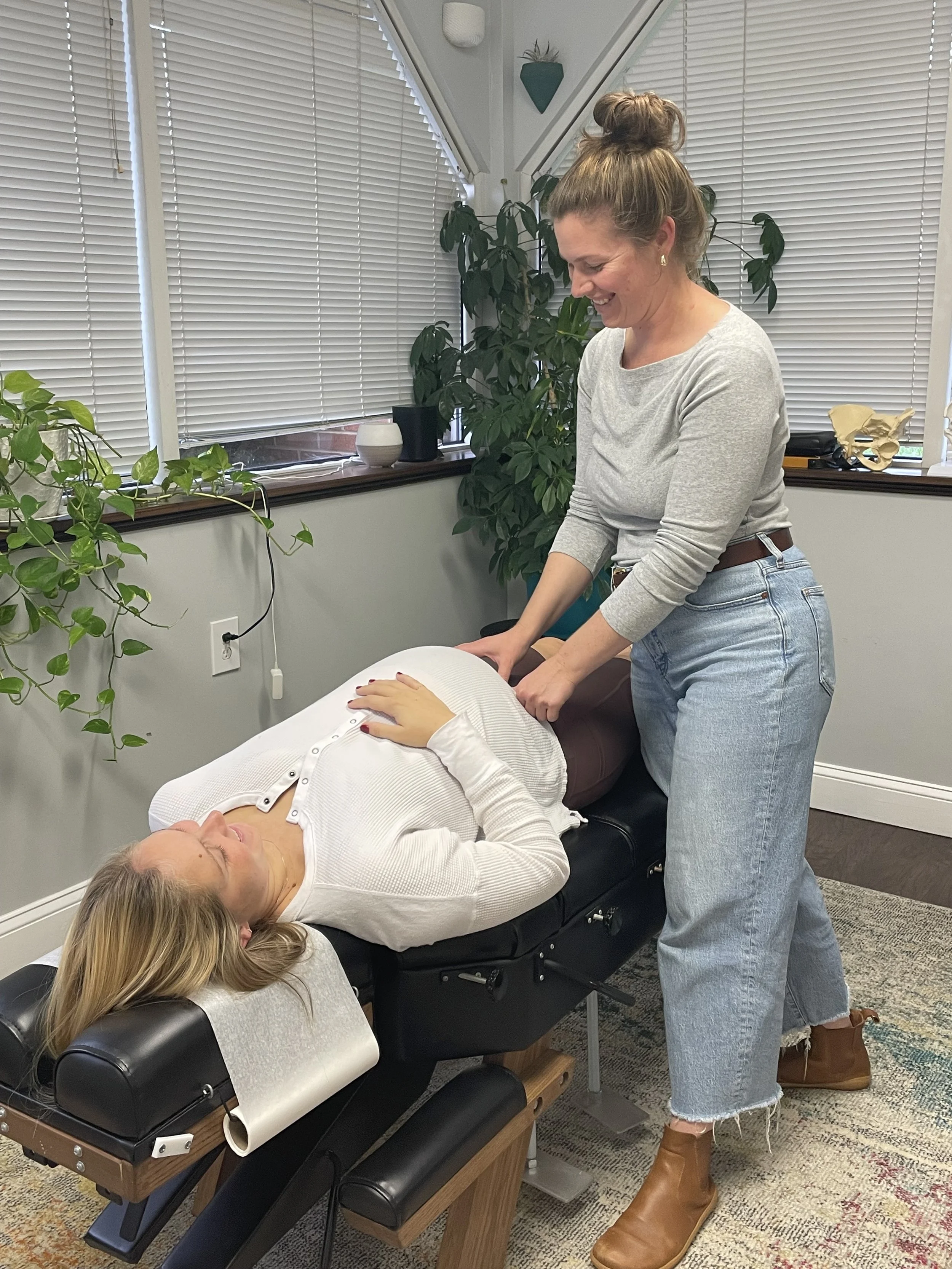 A woman receiving a massage from a massage therapist in a room with lots of green plants and window blinds.