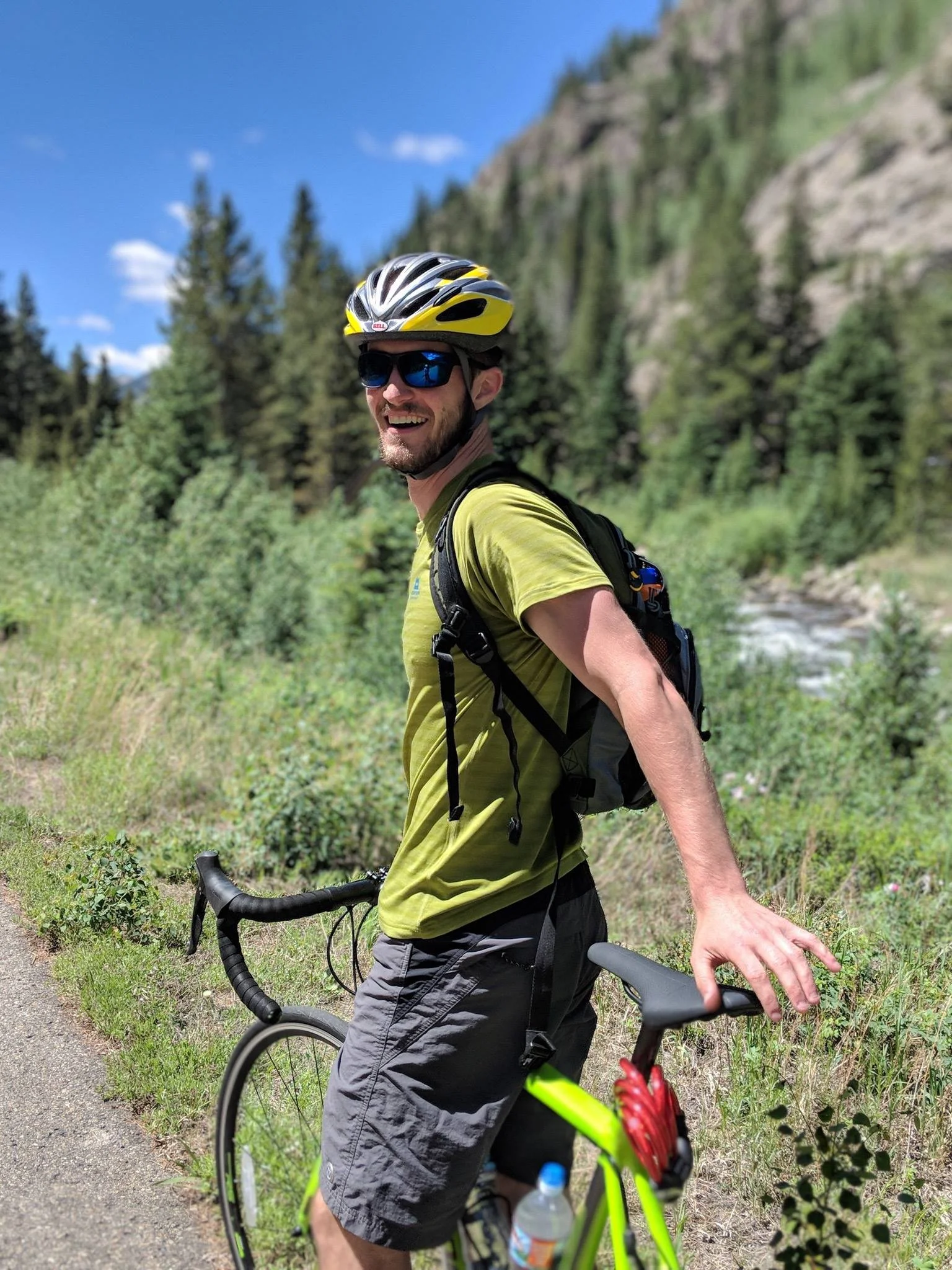 Man in yellow helmet and sunglasses smiling while holding a bicycle on a trail near a forested mountain landscape.