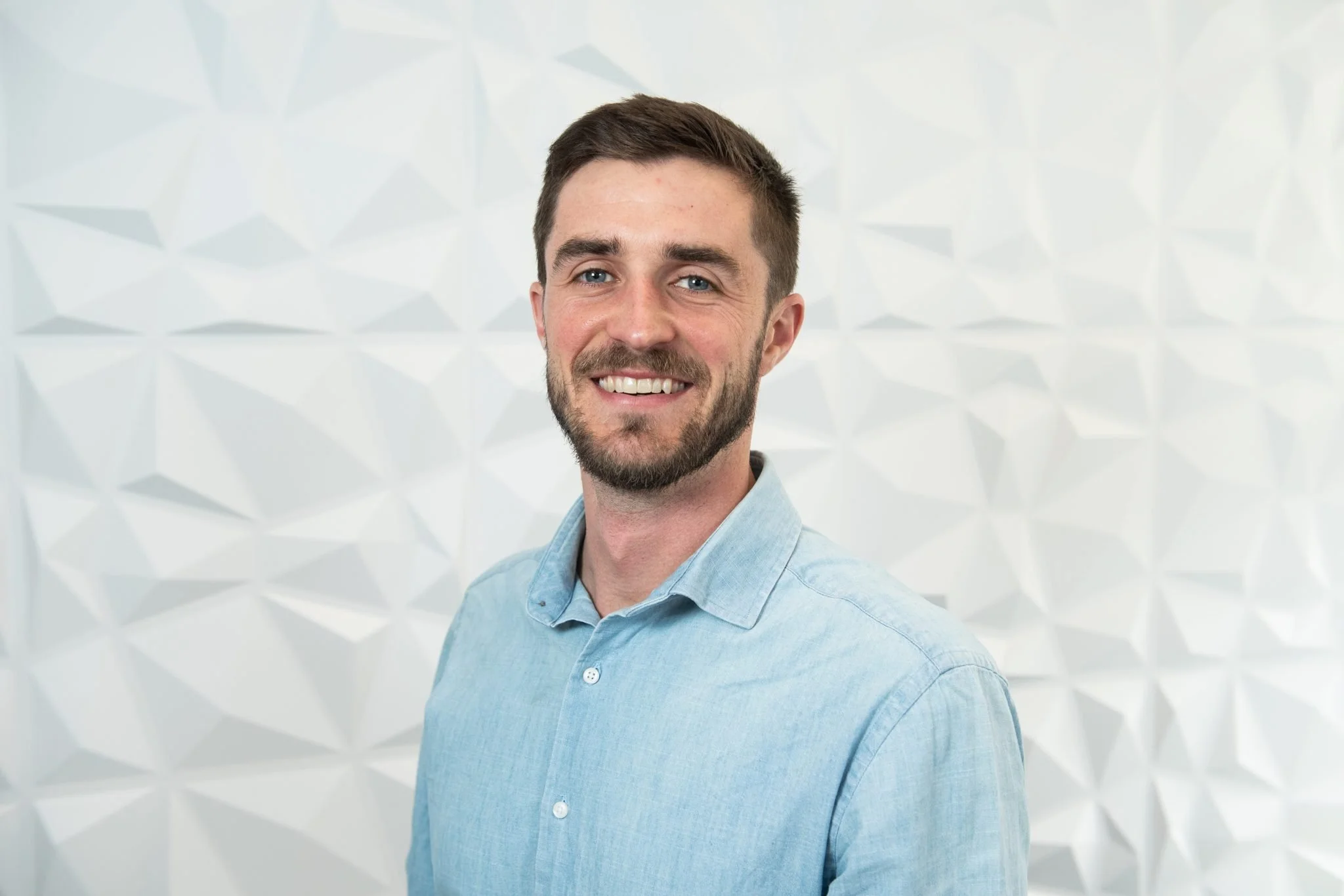 A man with brown hair, beard, and blue eyes smiling, wearing a light blue shirt, standing in front of a white geometric-patterned wall.