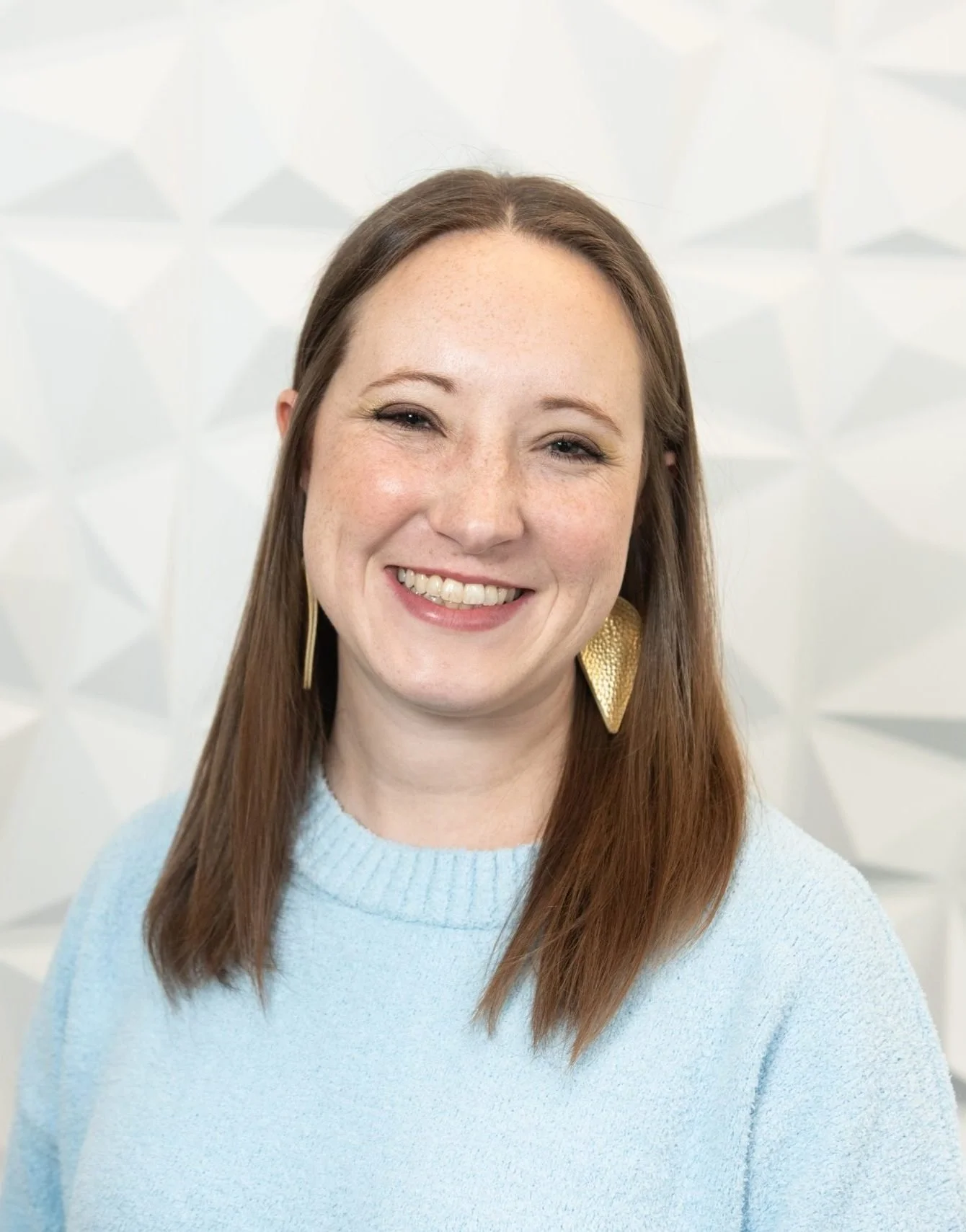 A woman with long brown hair, wearing a light blue sweater and gold teardrop-shaped earrings, smiling in front of a white background with geometric patterns.