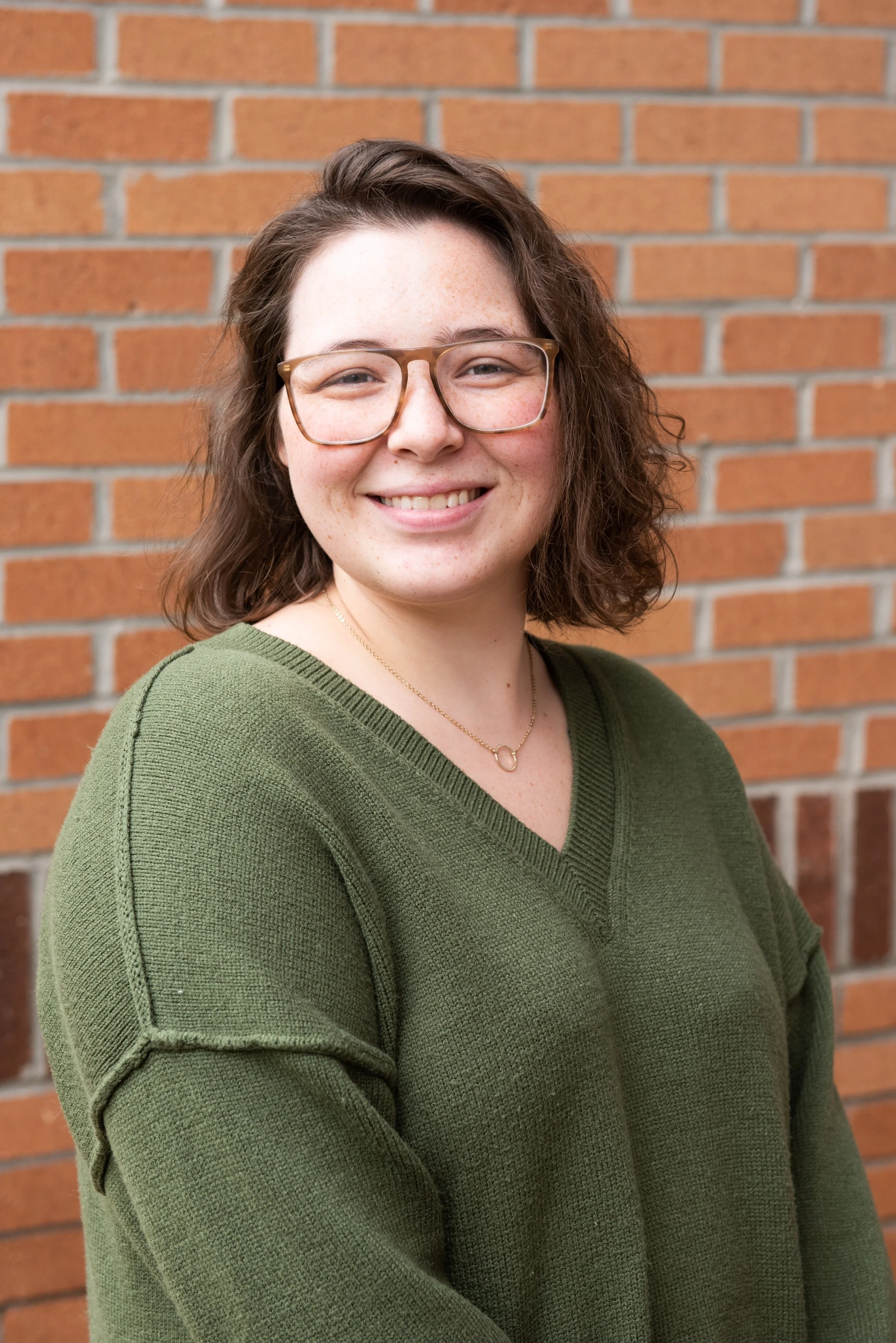 A woman with shoulder-length curly brown hair, wearing glasses and a green sweater, smiling in front of a brick wall.