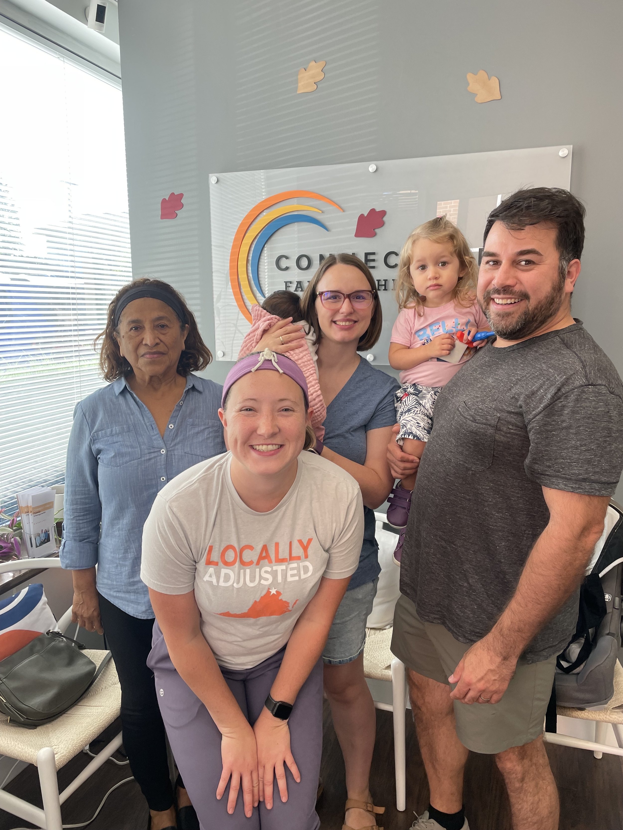 A group of six people, including children and adults, gathered indoors smiling for a photo. Behind them is a sign that reads 'Connect Family Health' with a colorful rainbow logo. The setting appears to be a medical or community health office.