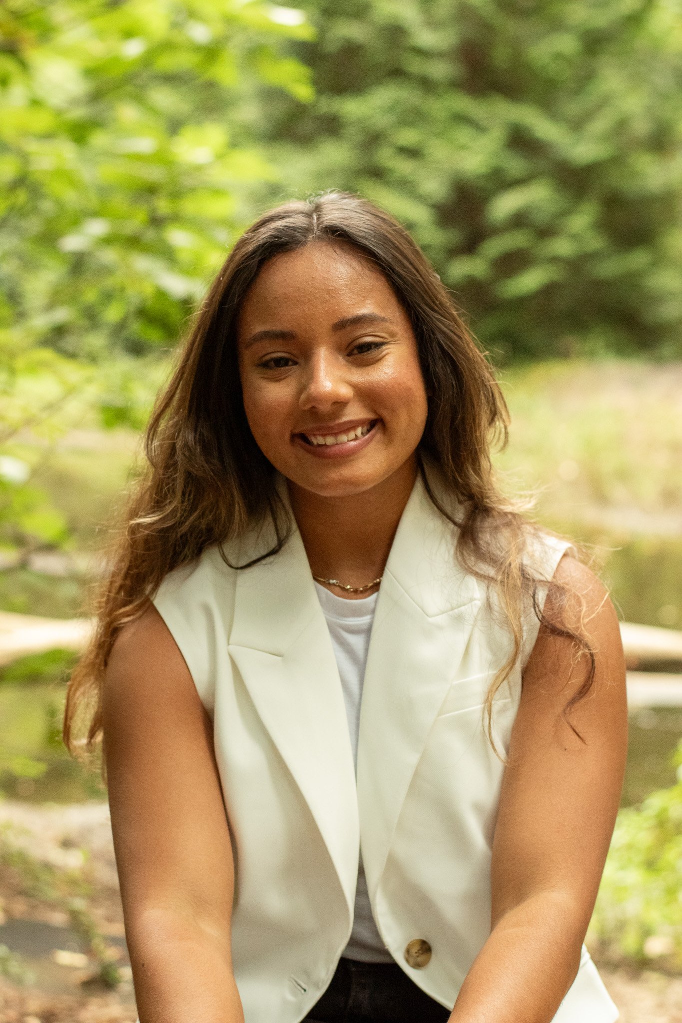 A woman with long, wavy brown hair smiling outdoors in a green, leafy environment. She is wearing a sleeveless white blazer over a white top, with a gold necklace.