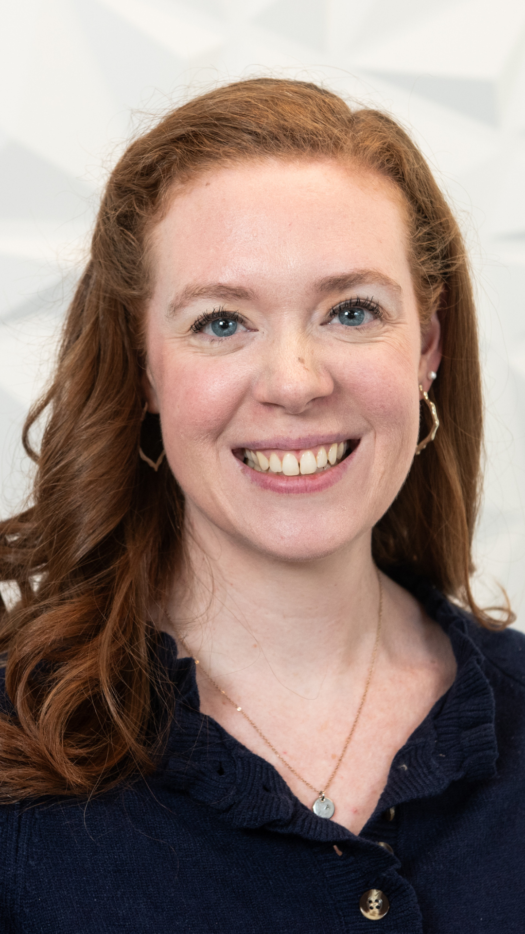 Close-up of a woman with red hair and blue eyes smiling, wearing pearl earrings and a dark blue knitted sweater.