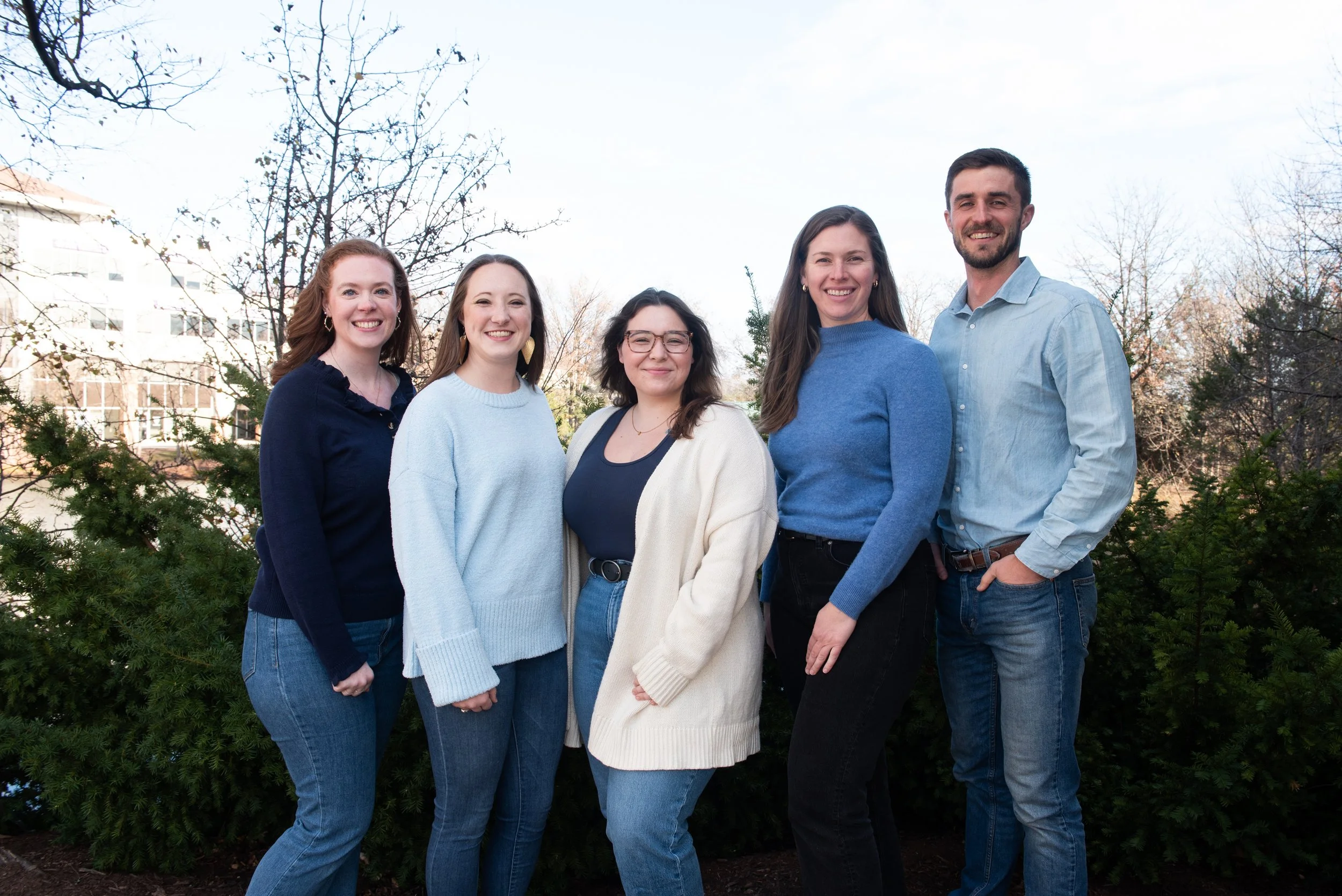 A group of five young adults, consisting of four women and one man, standing outdoors in front of trees and shrubs, smiling at the camera on a clear day.
