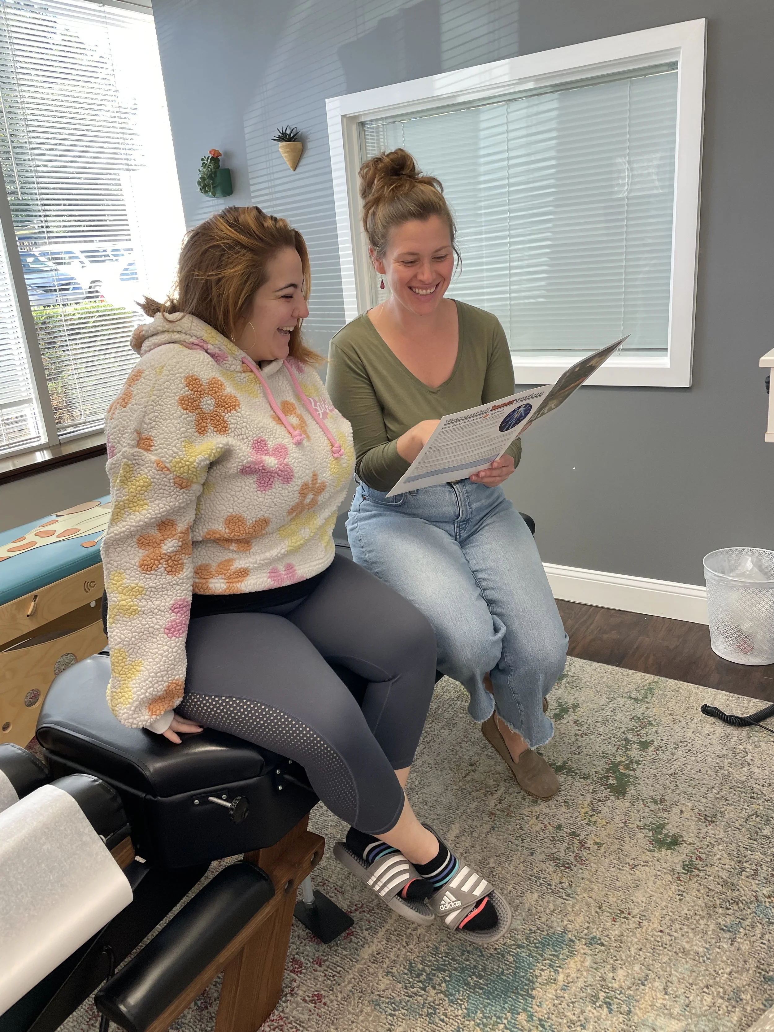 Two women sitting together indoors, looking at a brochure and smiling, with a window, grey walls, and a decorative wall plan in the background.