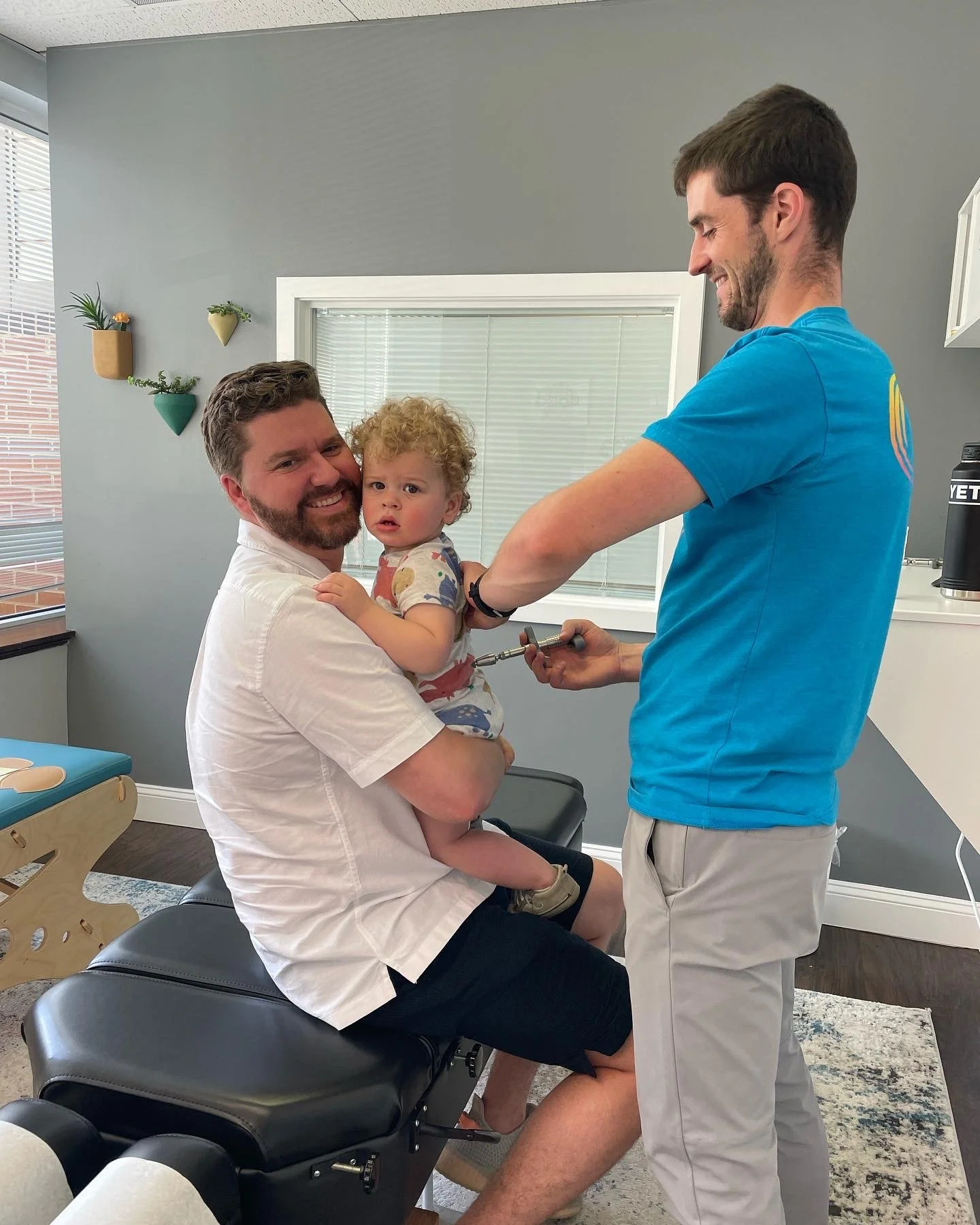 A man holding a young child on his lap at a medical clinic, with a friendly dentist or doctor preparing to examine the child's mouth, in a bright room with gray walls and potted plants.