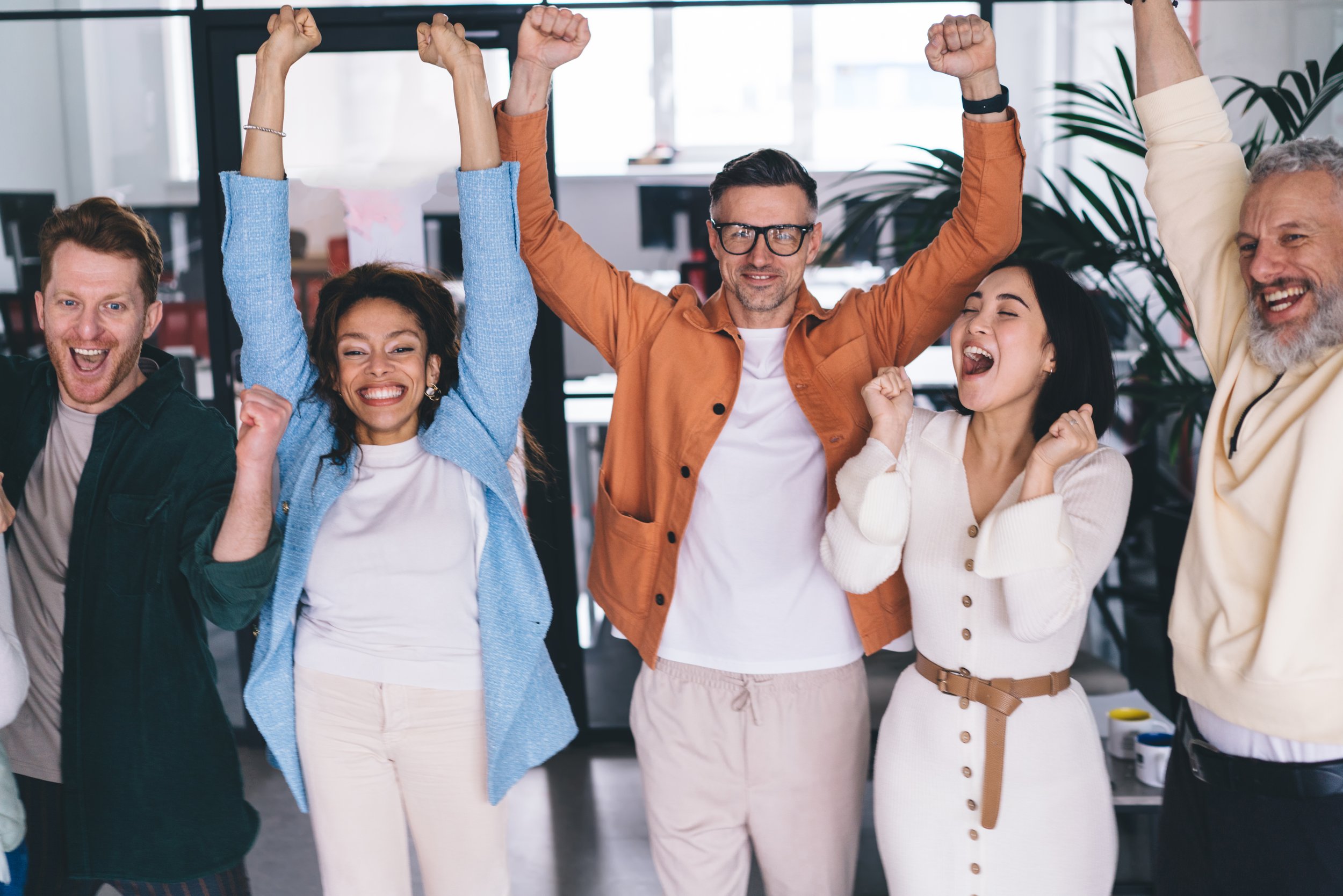 A diverse group of five people celebrating with raised fists and smiling, standing close together in a bright indoor office setting.