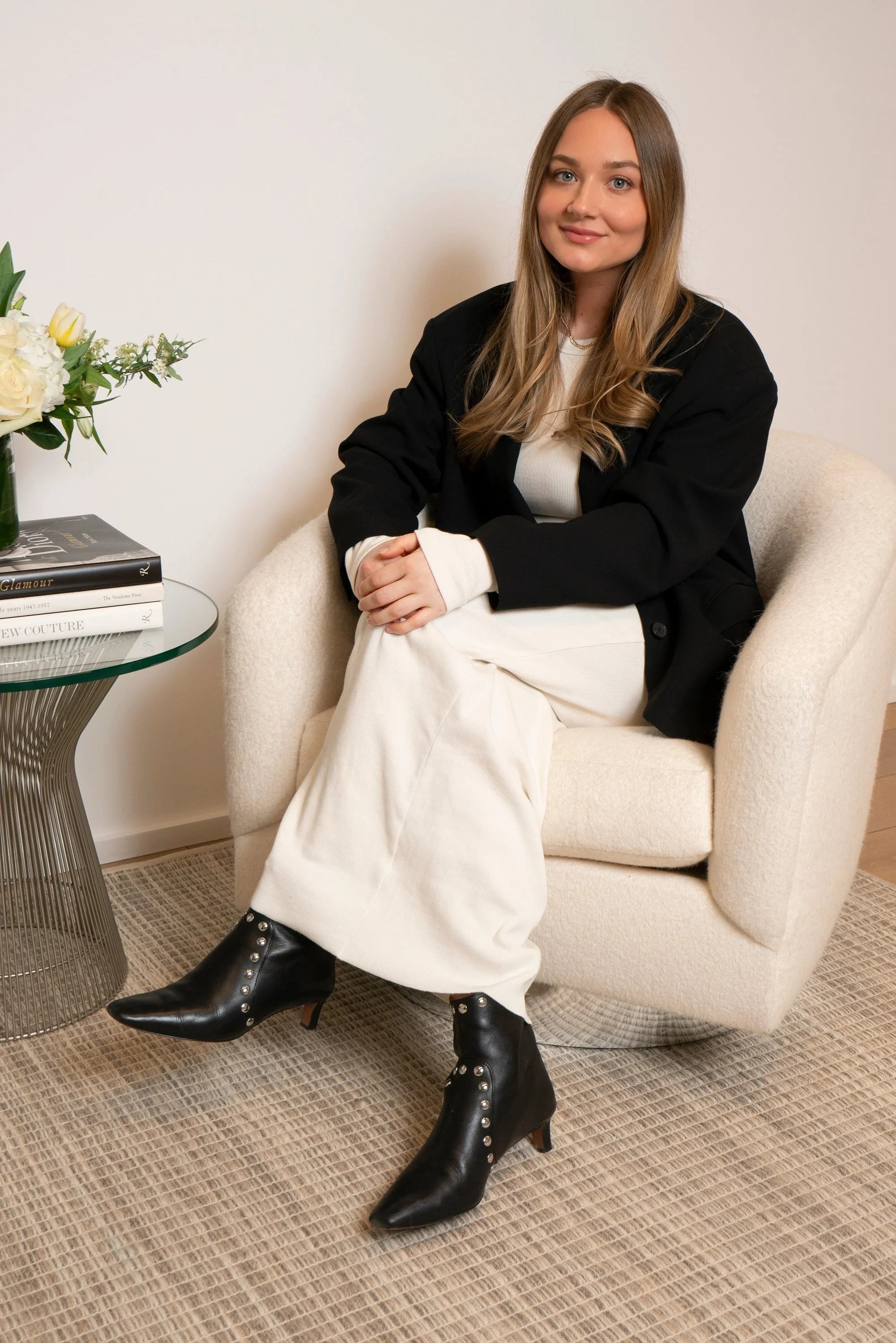 Lindsey Weisbrot, founder of LW Collective, seated in a cream armchair in a black blazer, white dress, and black boots.