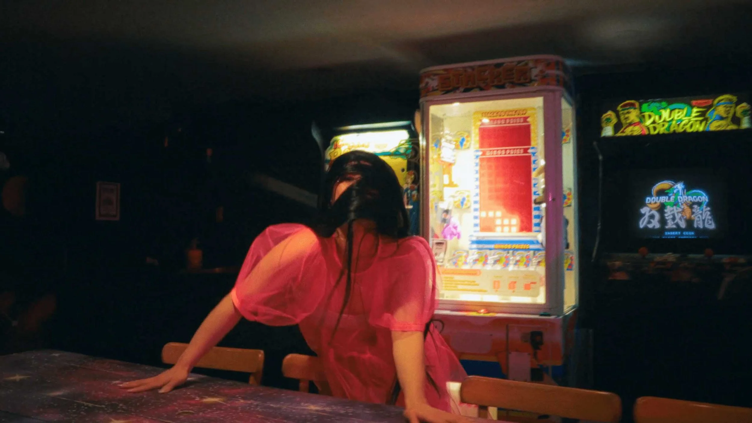 A still image from the short film Stone Skin. A person with long dark hair wearing a pink dress leaning over a table, with arcade machines in the background. 