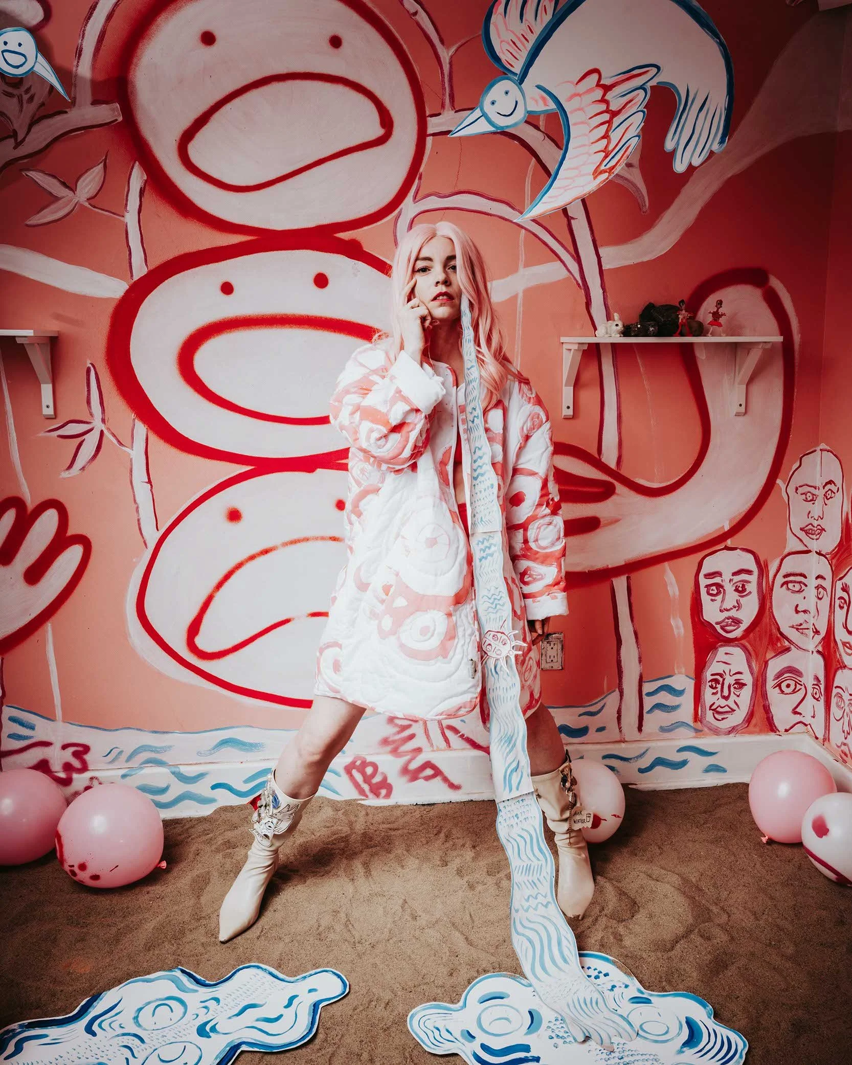 A portrait of artist Rachel Gray. She wears a pink wig and a painted coat. A paper tear streams from her eye to the floor. There are painted cartoon faces on the walls.
