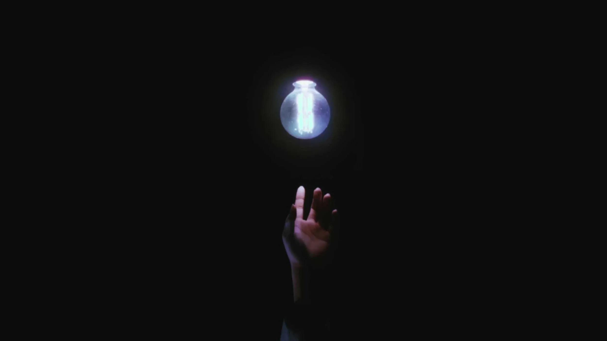 A still image from the short film Stone Skin. A hand reaching towards a glowing light bulb in a dark environment.