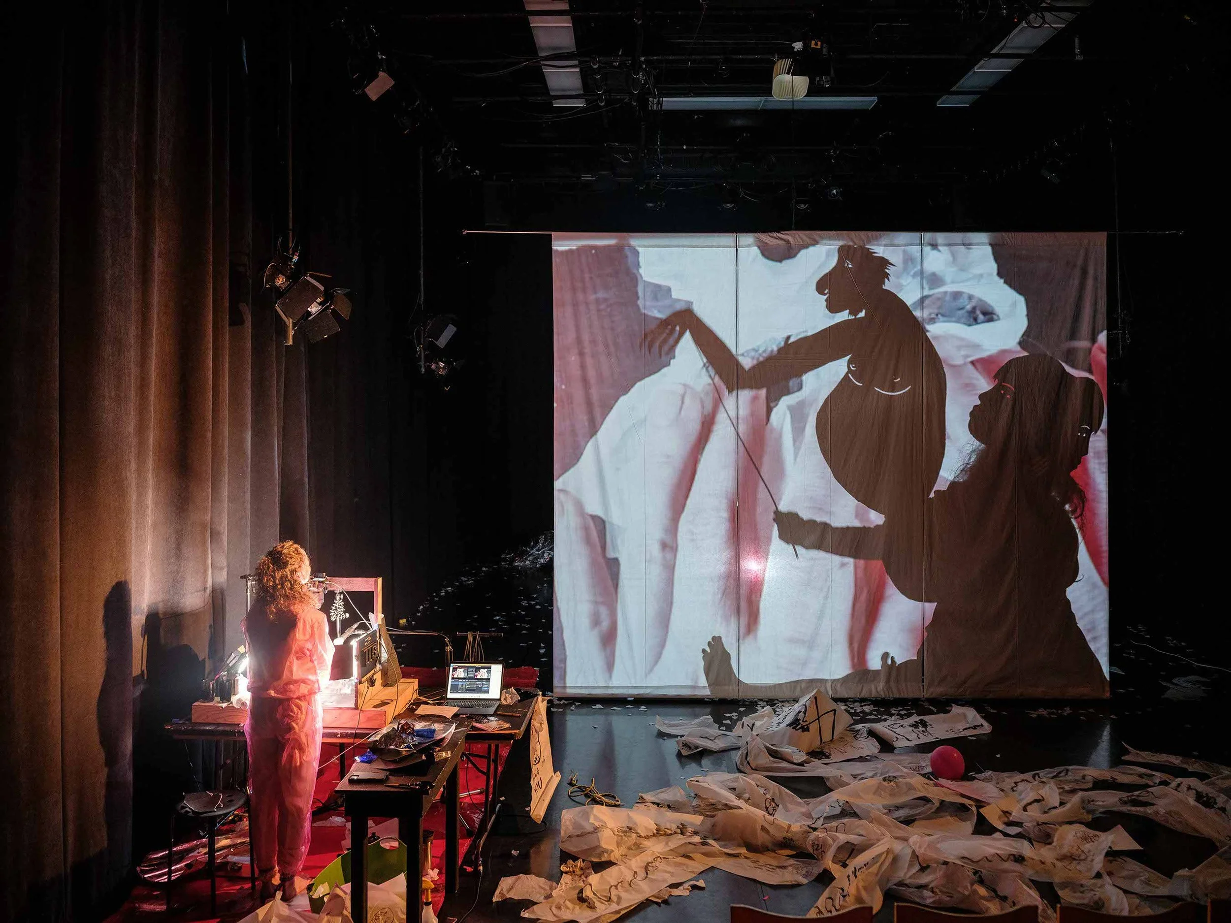 Behind a screen a figure in a mask is holding a shadow puppet monkey. Rachel Gray stands in front of the screen drawing. 