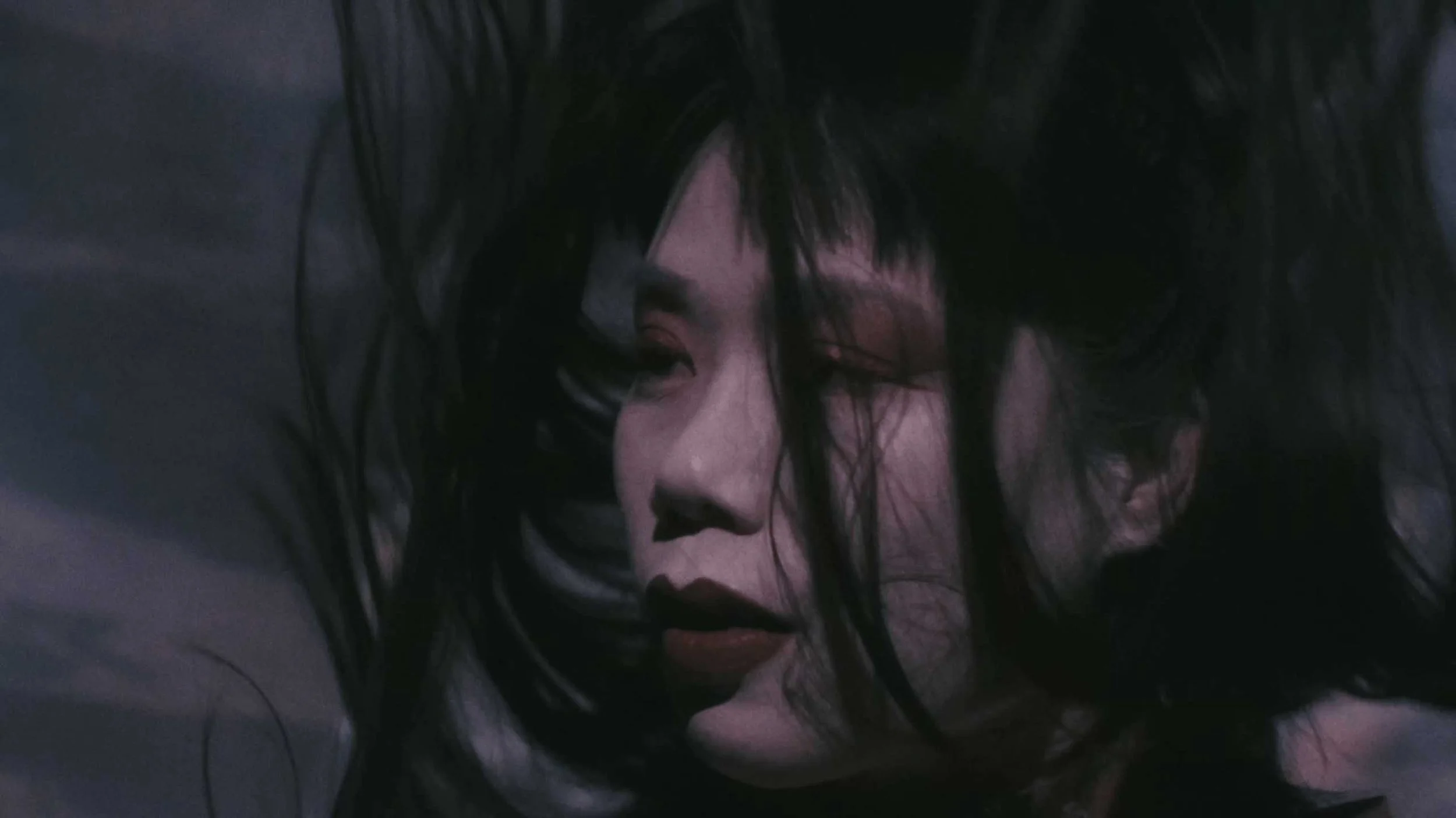 A still image from the short film Stone Skin. Close-up of a person with long dark hair and makeup, looking down with a serious expression.