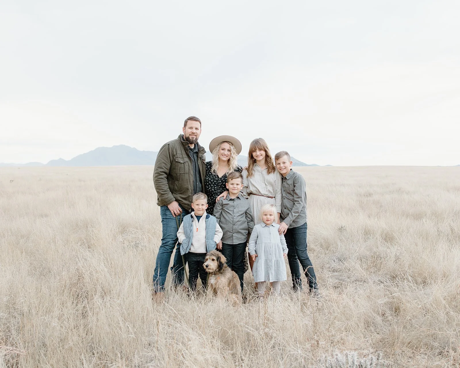 Family of eight people and a dog standing in a grassy field with mountains in the background.