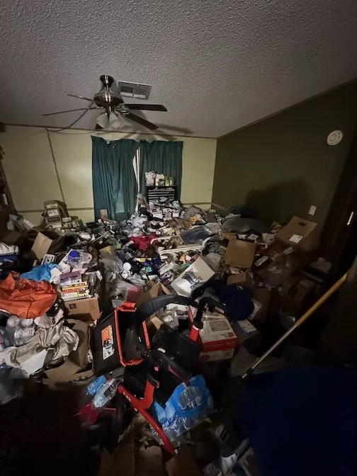 Messy living room filled with clutter, boxes, and scattered items, with a ceiling fan and teal curtains in the background.