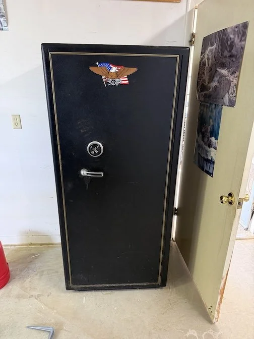 A black gun safe with a Presidential Seal sticker on the top center, positioned in a room with a white wall and an open door to the right with pictures taped to it.