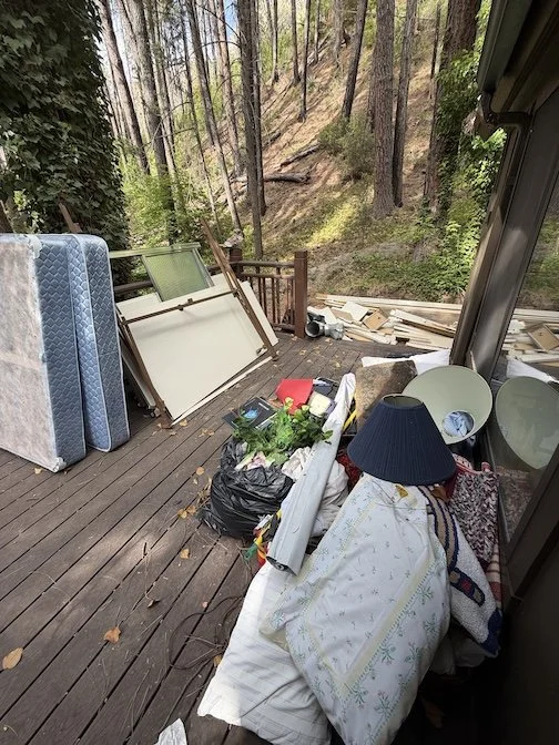 Disorganized clutter outside on a wooden deck, including a mattress, picture frames, a black trash bag filled with items, a lamp with a blue shade, and a pile of lumber, with a wooded hillside in the background.