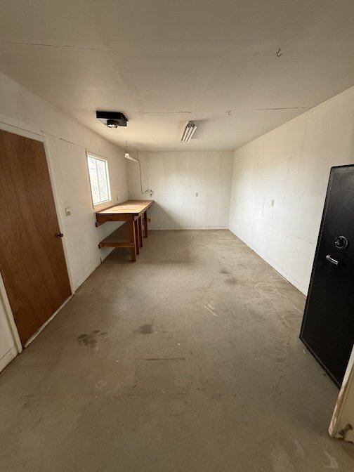 Empty room with a concrete floor, white walls, partially visible wooden door, window, wooden table with a shelf underneath, a ceiling light, and a black safe.