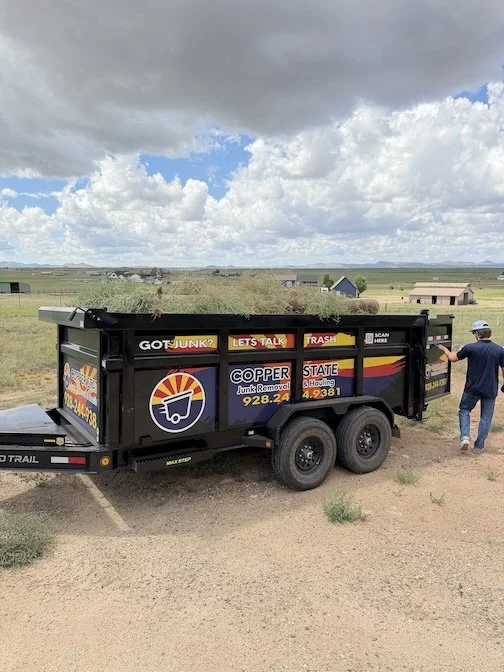 A black junk removal trailer with colorful advertising logos and text is parked in a rural area on dirt ground. The trailer has a pile of green yard debris inside, and a man in a blue shirt and hat is walking beside it, looking into the trailer. The background features open fields, a few small structures, and a partly cloudy sky.
