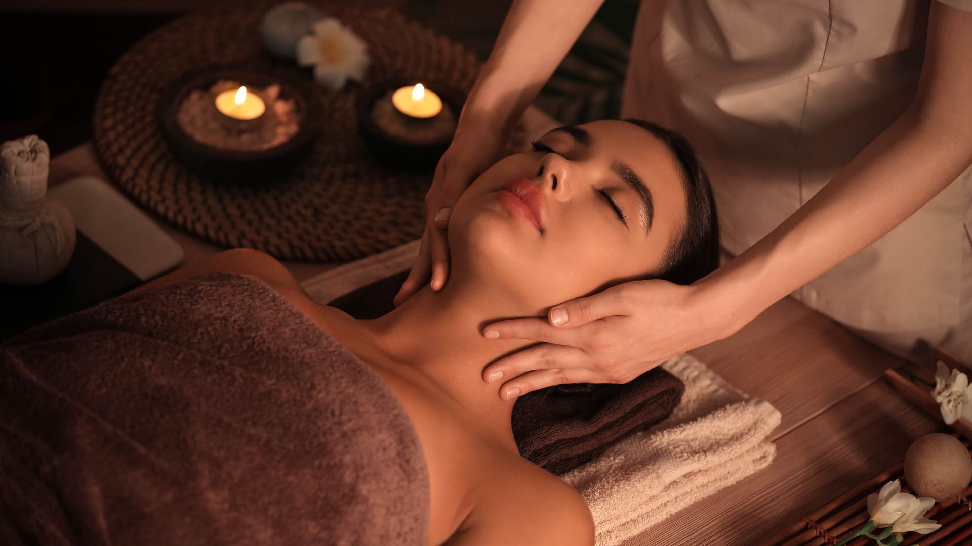 A woman receives a facial massage at a spa, lying on a massage table with her eyes closed, covered with a towel, and a relaxing ambiance created by candles and flowers in the background.