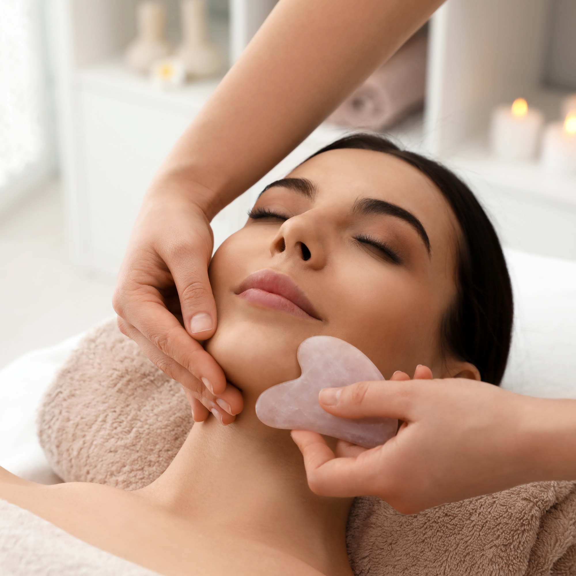 A woman lying down with closed eyes, being gently touched on her face and neck by a person's hands. The background features candles and a bright, softly lit room.