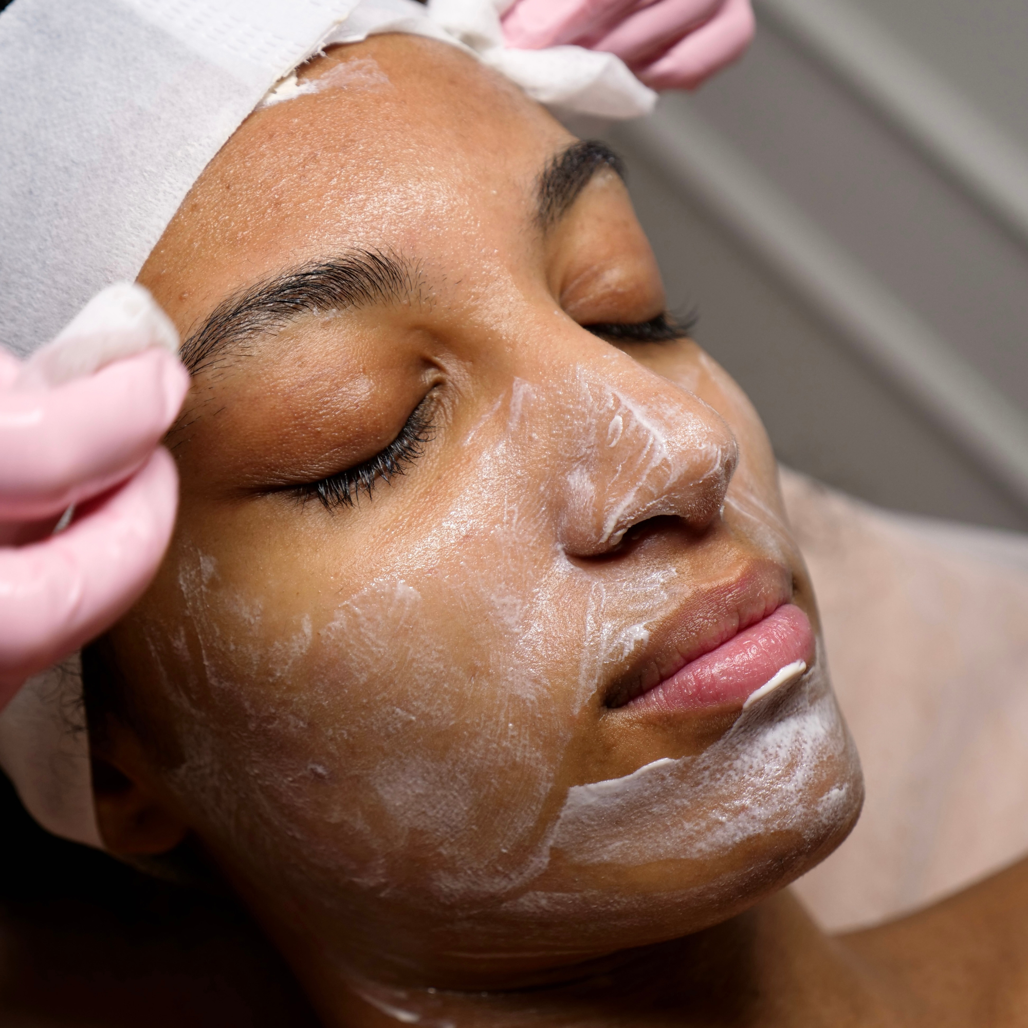 Close-up of a person receiving a facial treatment, lying down with eyes closed, with a facial mask applied to their face and a healthcare professional wearing pink gloves attending to them.
