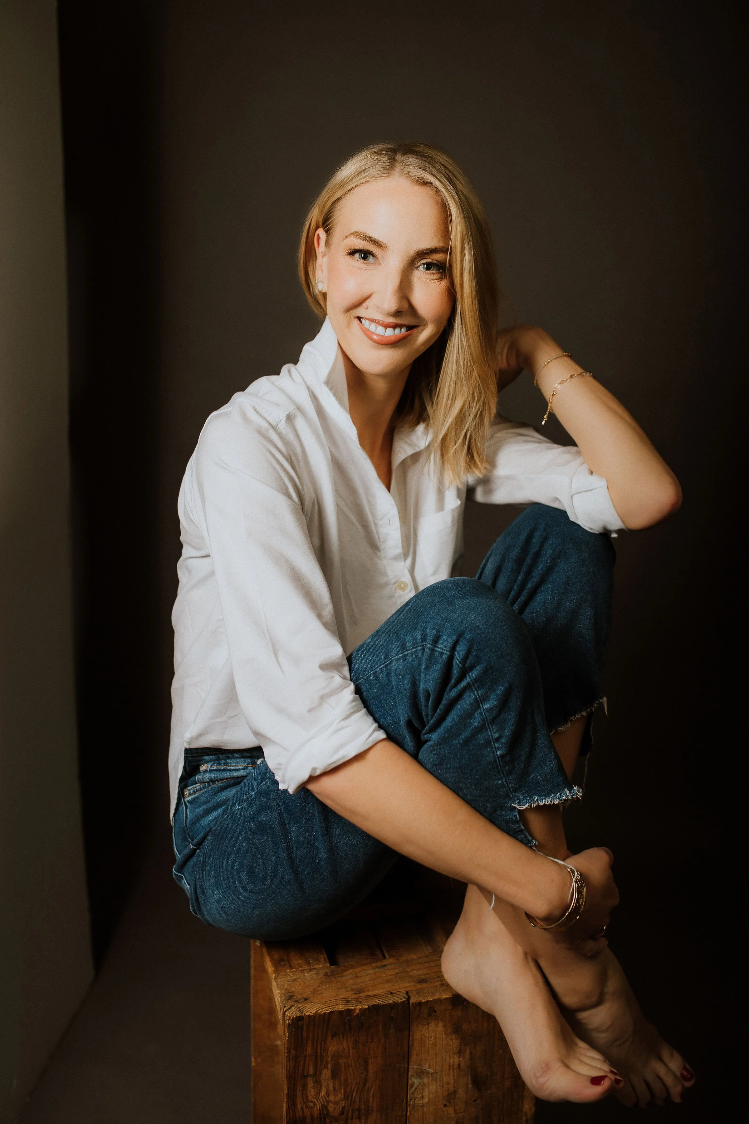 A woman with blonde hair, wearing a white shirt and blue jeans, sitting on a wooden block with her legs crossed. She is smiling, looking at the camera, and has jewelry on her right wrist and left ear. The background is dark.