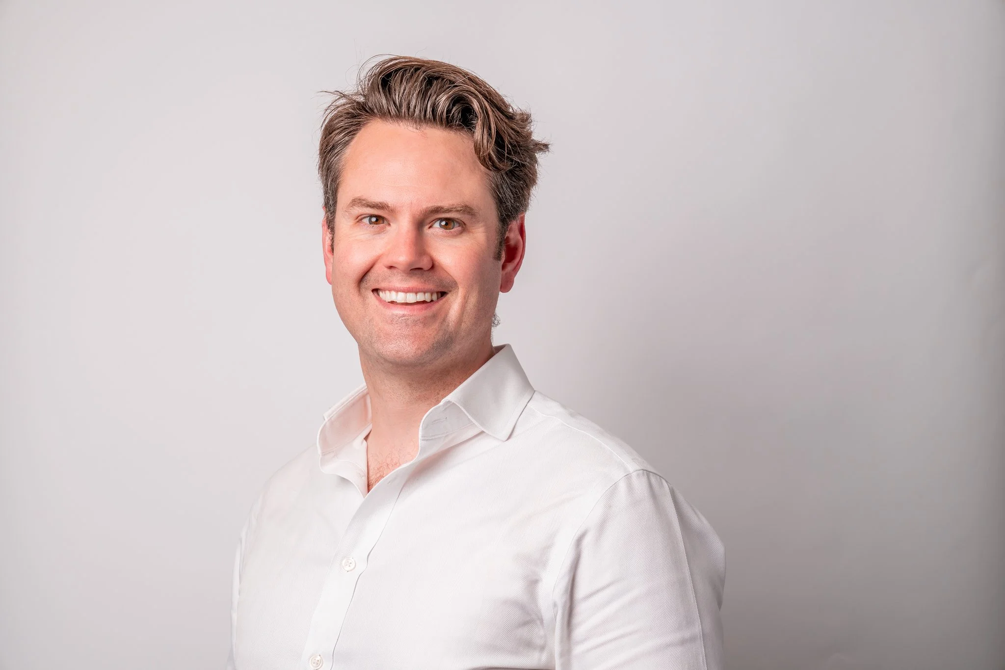 A smiling man with light brown hair wearing a white button-up shirt posing against a plain light gray background.