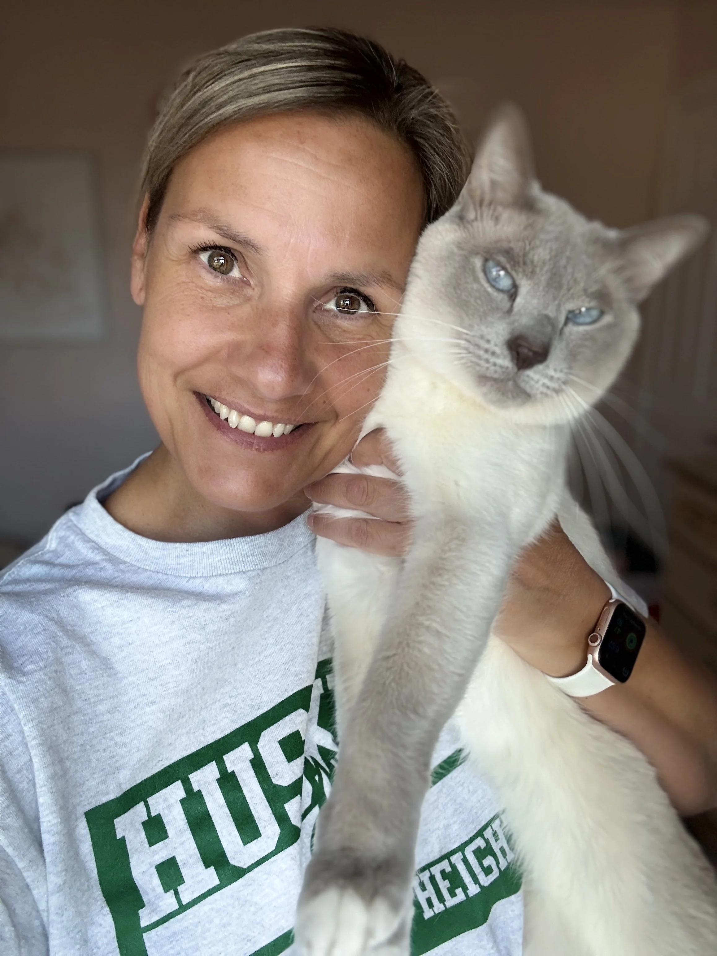A woman smiling while holding a gray cat with blue eyes close to her face.