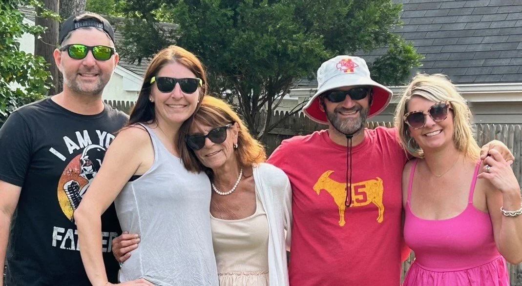 A group of five adults smiling in a backyard, dressed casually, with trees and a house in the background.