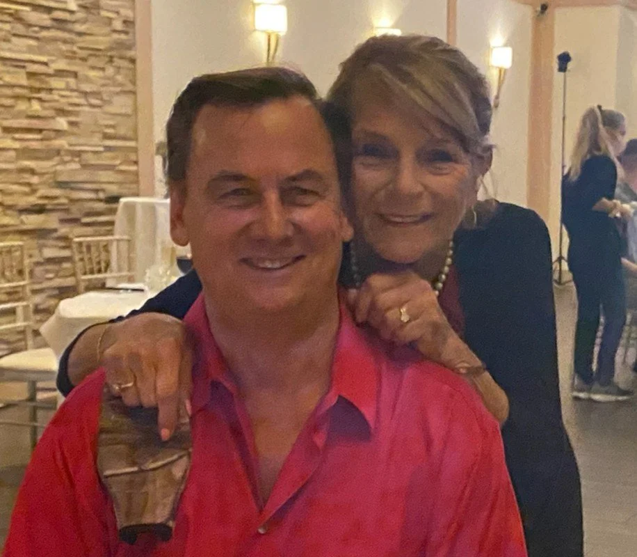 A man and Pamela Kossick Gant smiling together, the woman standing behind and leaning on the man's shoulders in a warmly lit indoor setting.