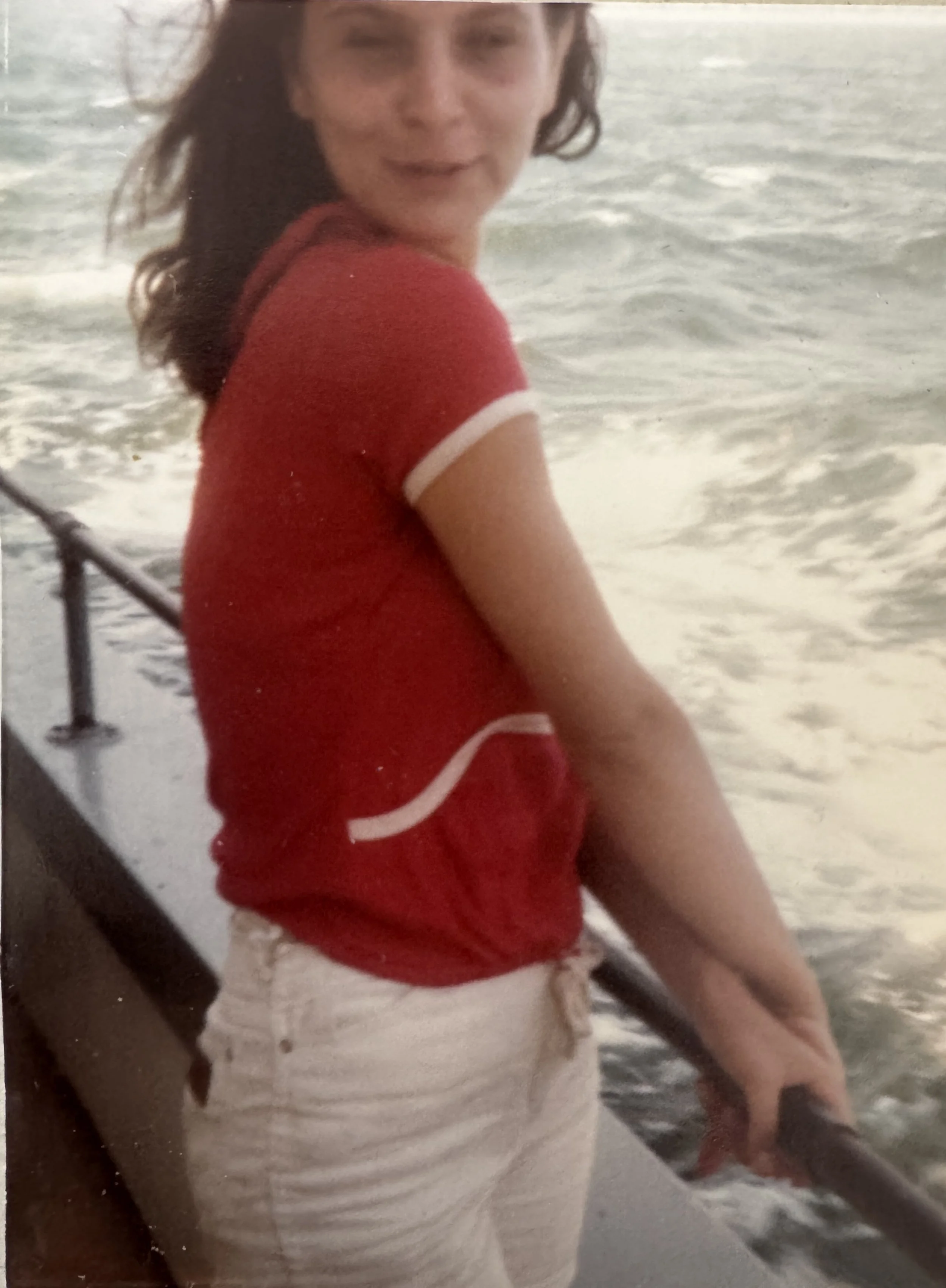 A woman in a red shirt and white shorts on a beach, holding a fish. The ocean is in the background.