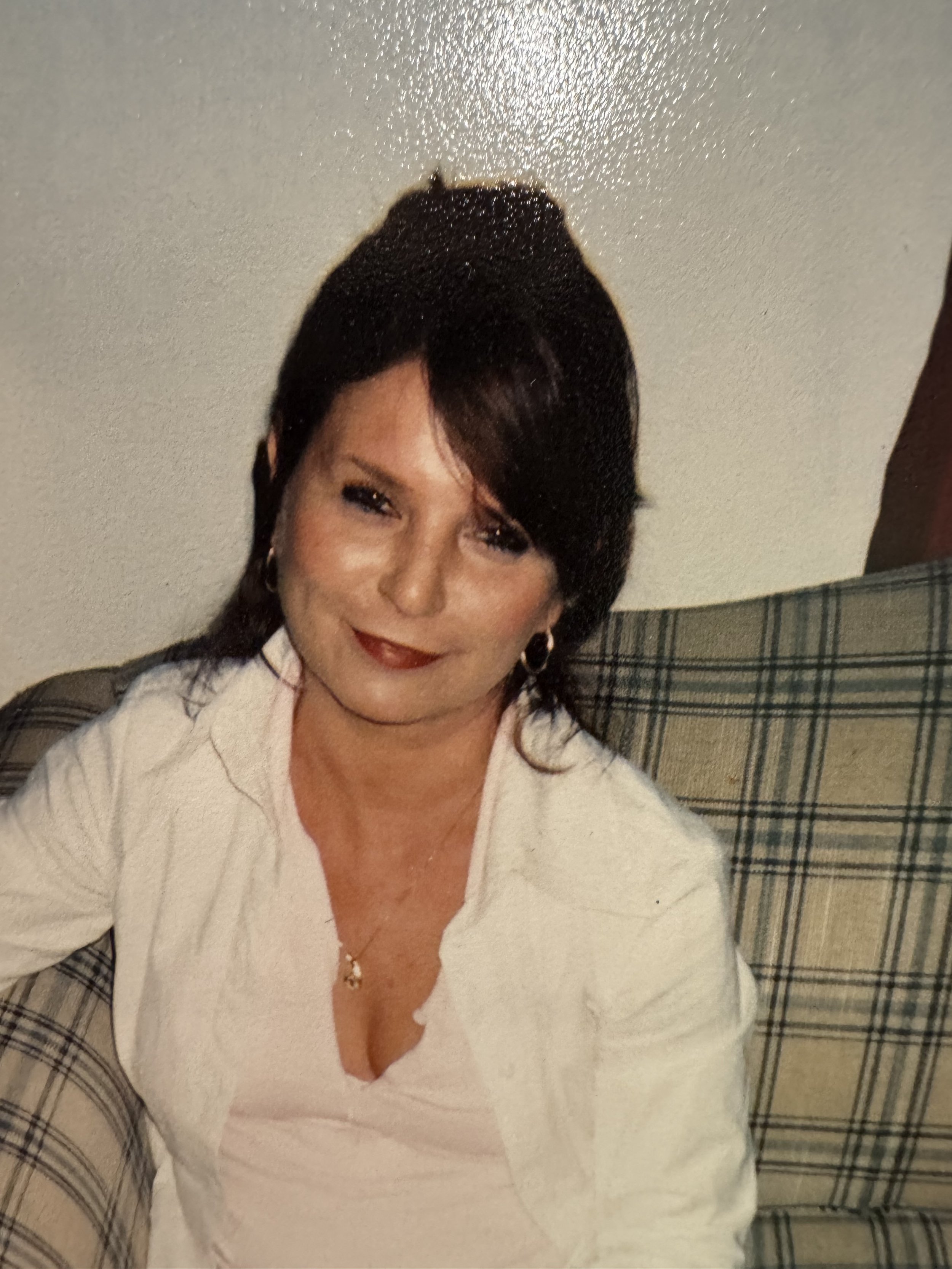 A woman with short dark hair, light makeup, and red lipstick sitting on a plaid upholstered piece of furniture, smiling slightly, wearing a cream-colored jacket over a white top, with a gold necklace and earrings.