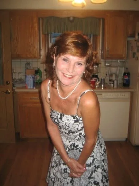 Pamela Kossick Gant with short red hair wearing a floral dress and pearl necklace in a kitchen, smiling at the camera.