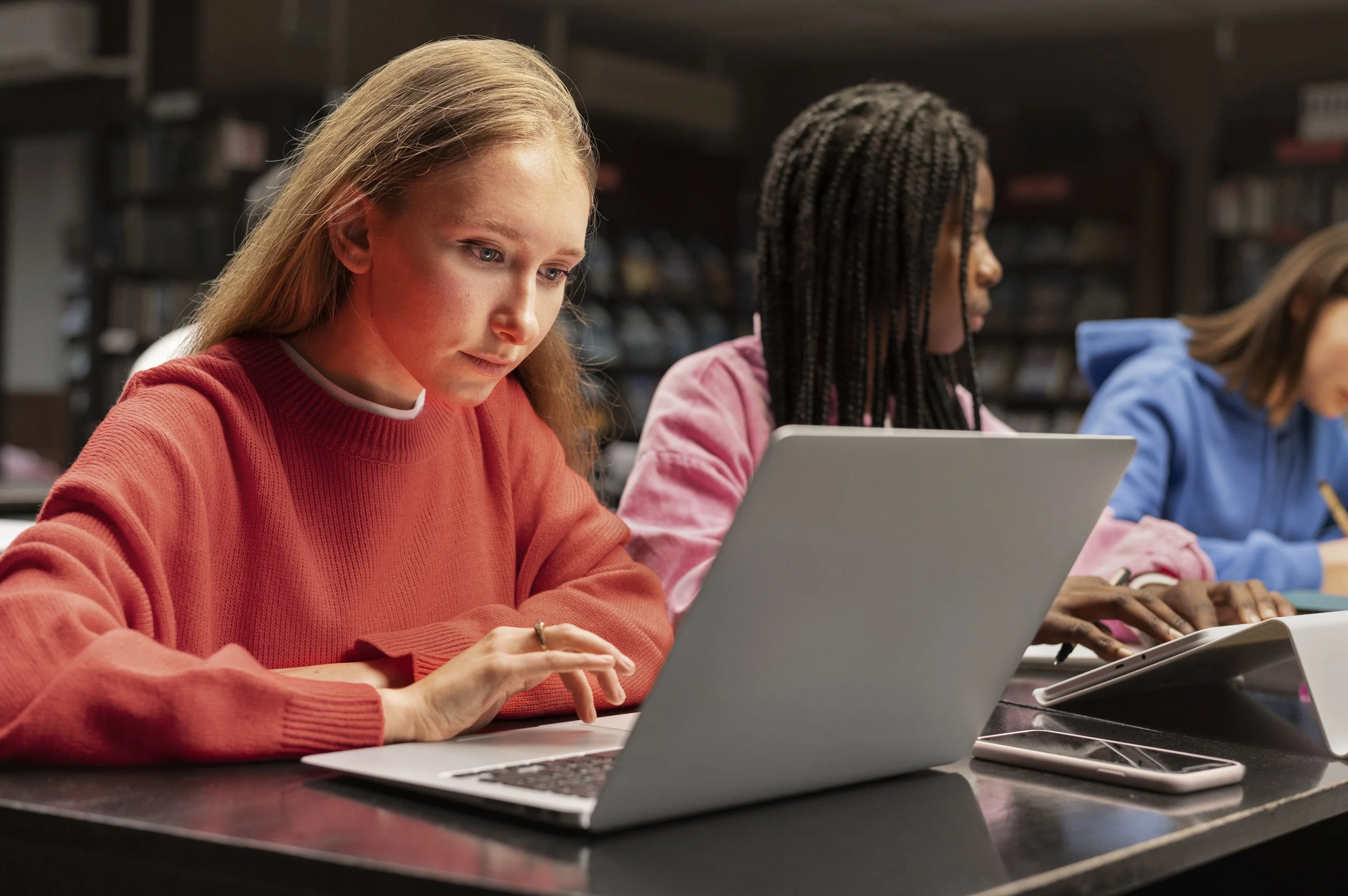 Students sitting at desks in a classroom, working on laptops and tablets, with a library in the background.